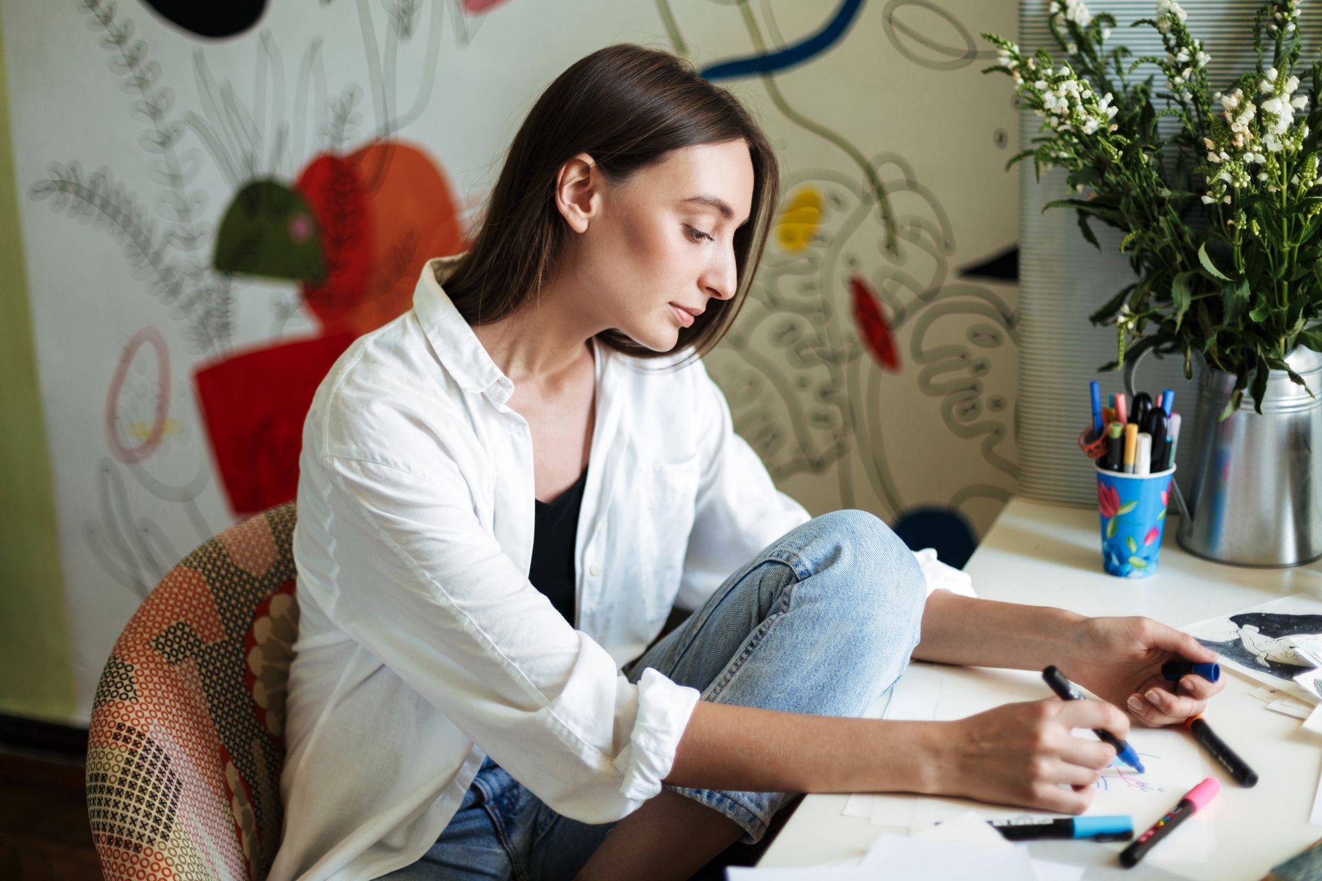 Woman in a white shirt sketching at a desk beside markers, a phone, and a plant in a bright room