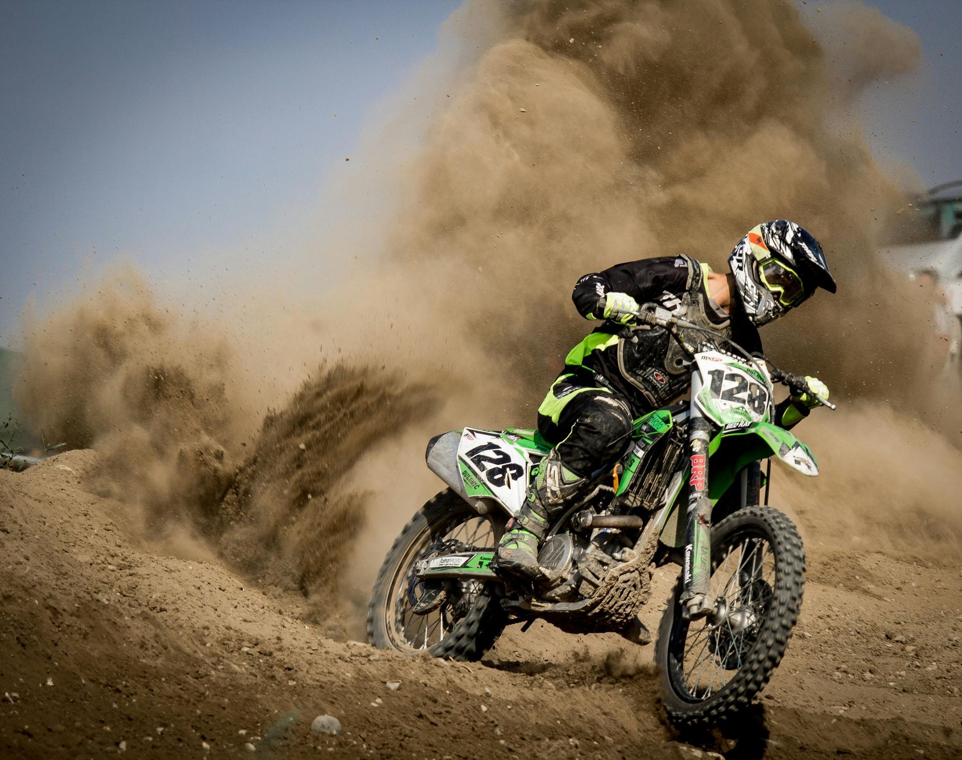 A motocross rider in green and black gear leans into a turn, kicking up a large cloud of dust on a dirt track.