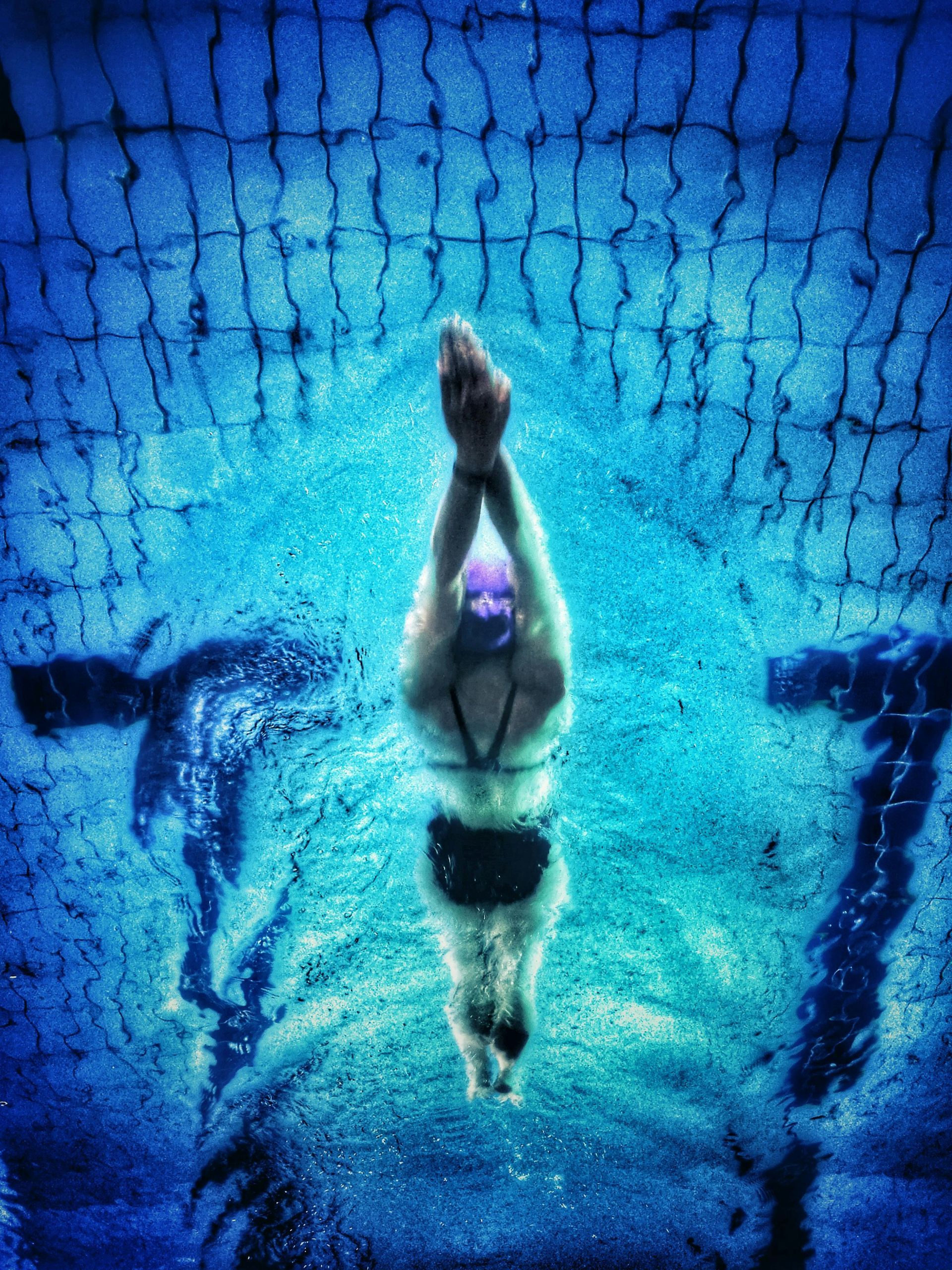 A swimmer underwater in a streamlined position, seen from above, gliding through a bright blue pool with lane markings.