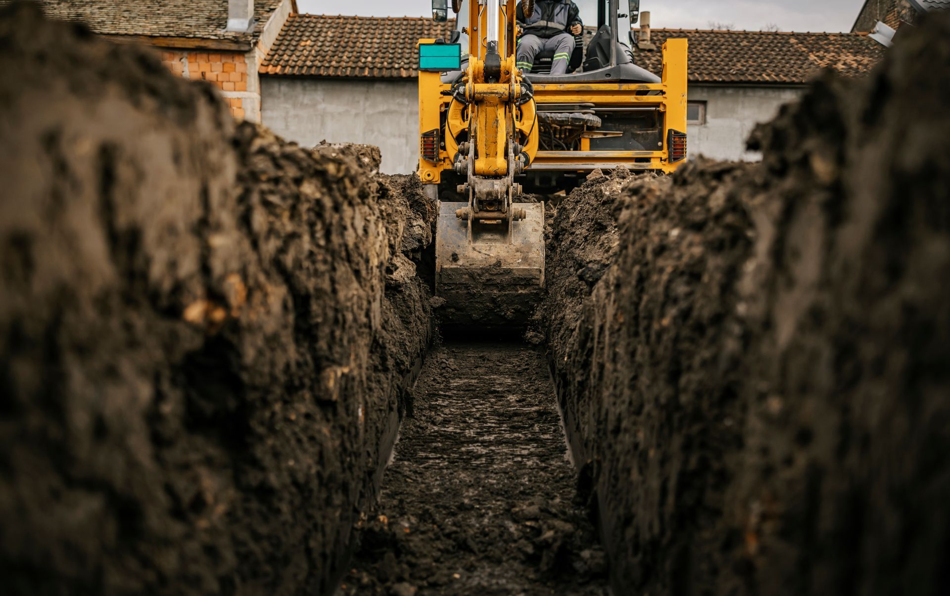 Excavator digging a trench between soil walls near residential buildings.