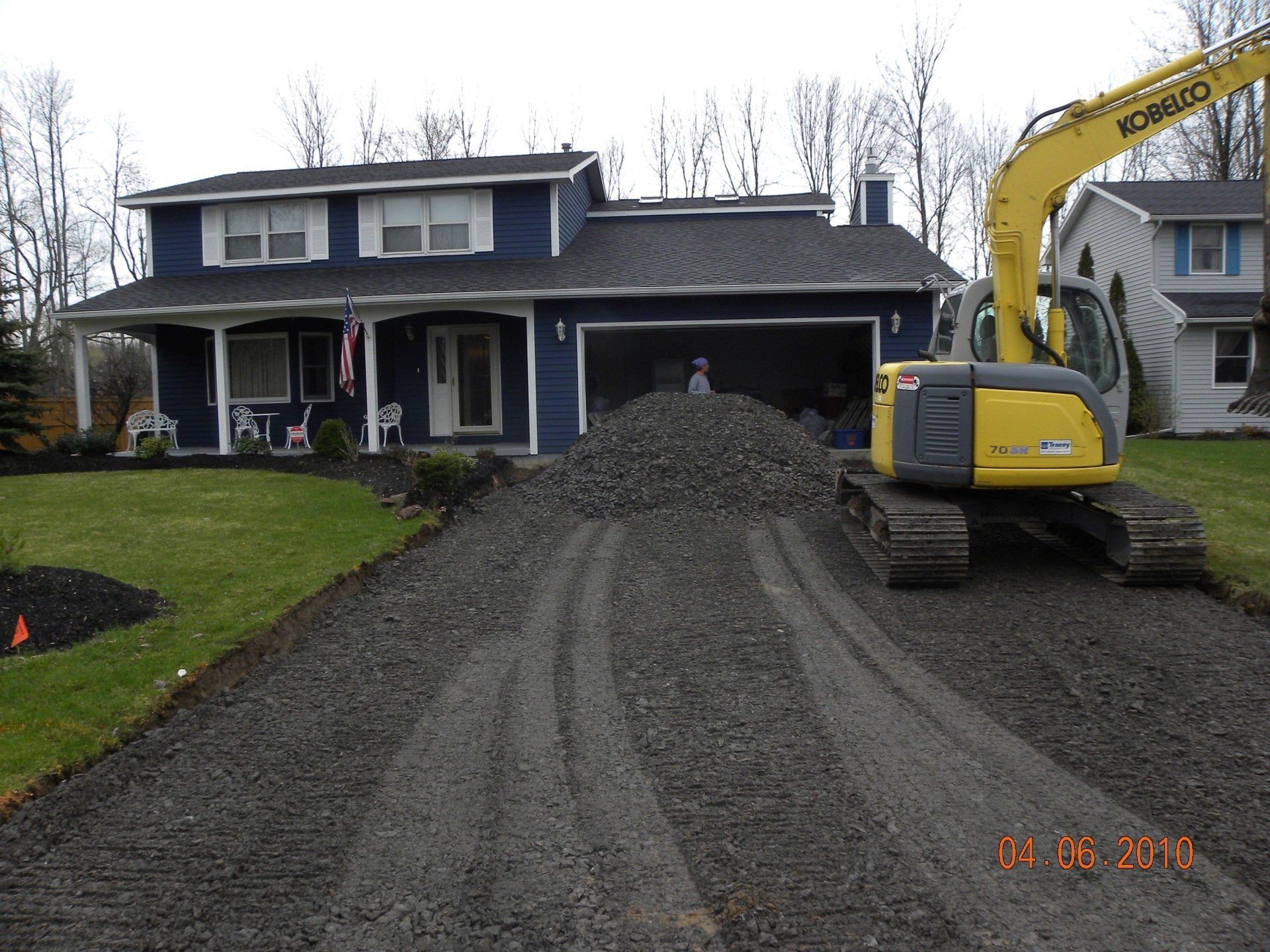 Excavator laying gravel on a driveway in front of a blue two-story house.