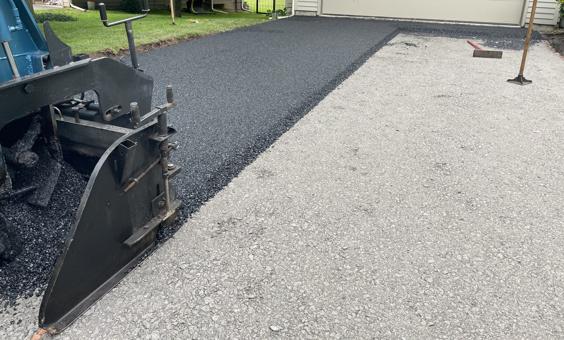 Asphalt paving machine laying blacktop on a driveway, next to a gravel surface.