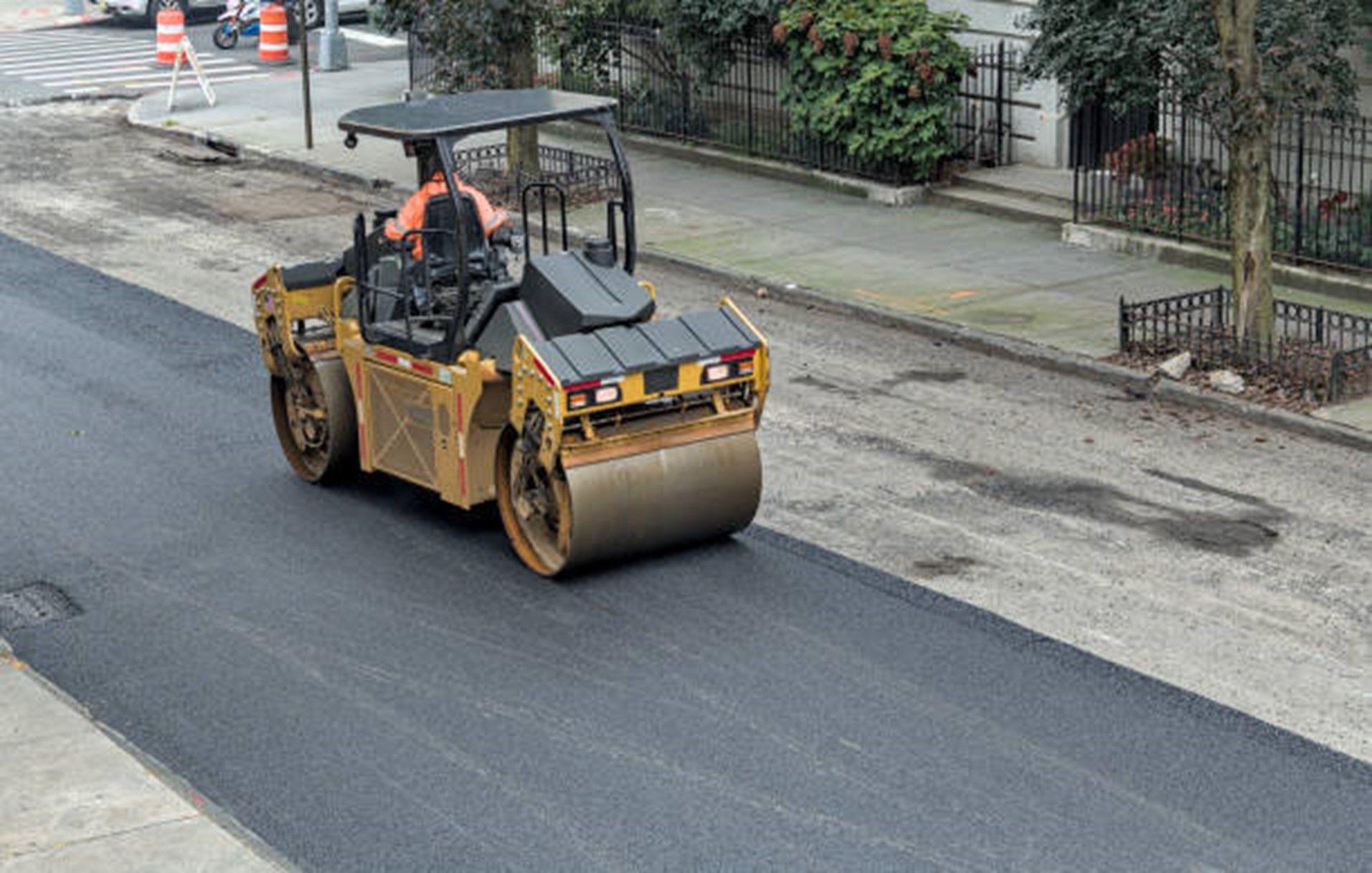 Road roller compacting fresh asphalt on a city street, operated by a worker wearing an orange safety vest. Road roller compacting fresh asphalt on a city street, operated by a worker wearing an orange safety vest.