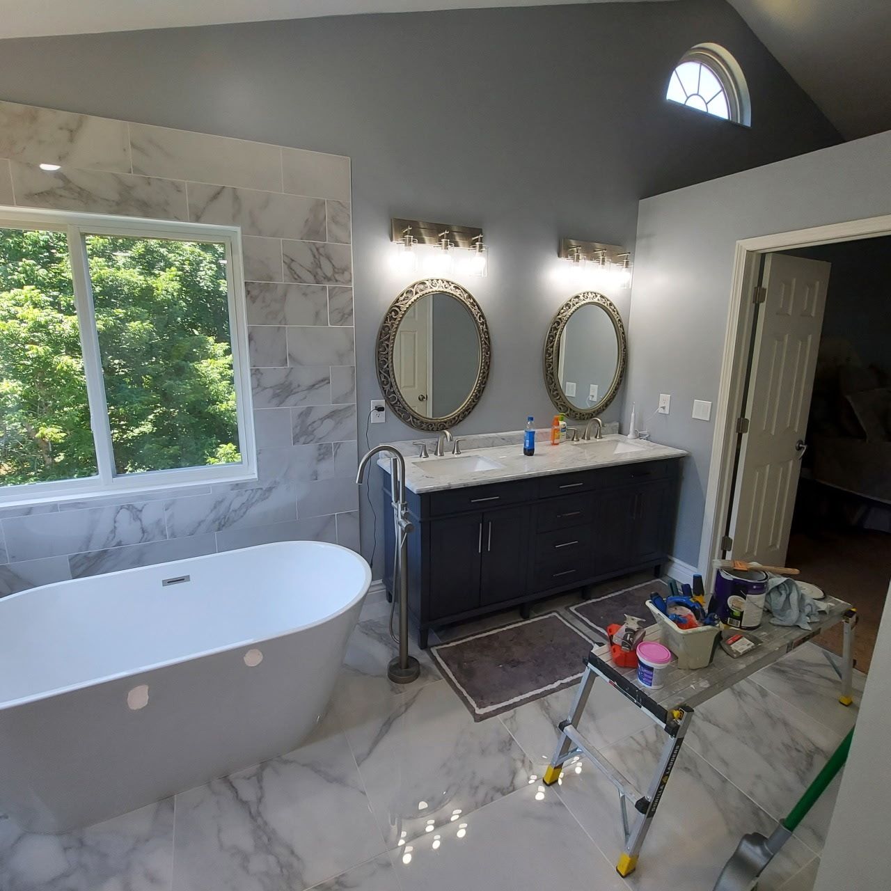 Bathroom with a white tub, marble tile, gray walls, double sinks, and a large window.