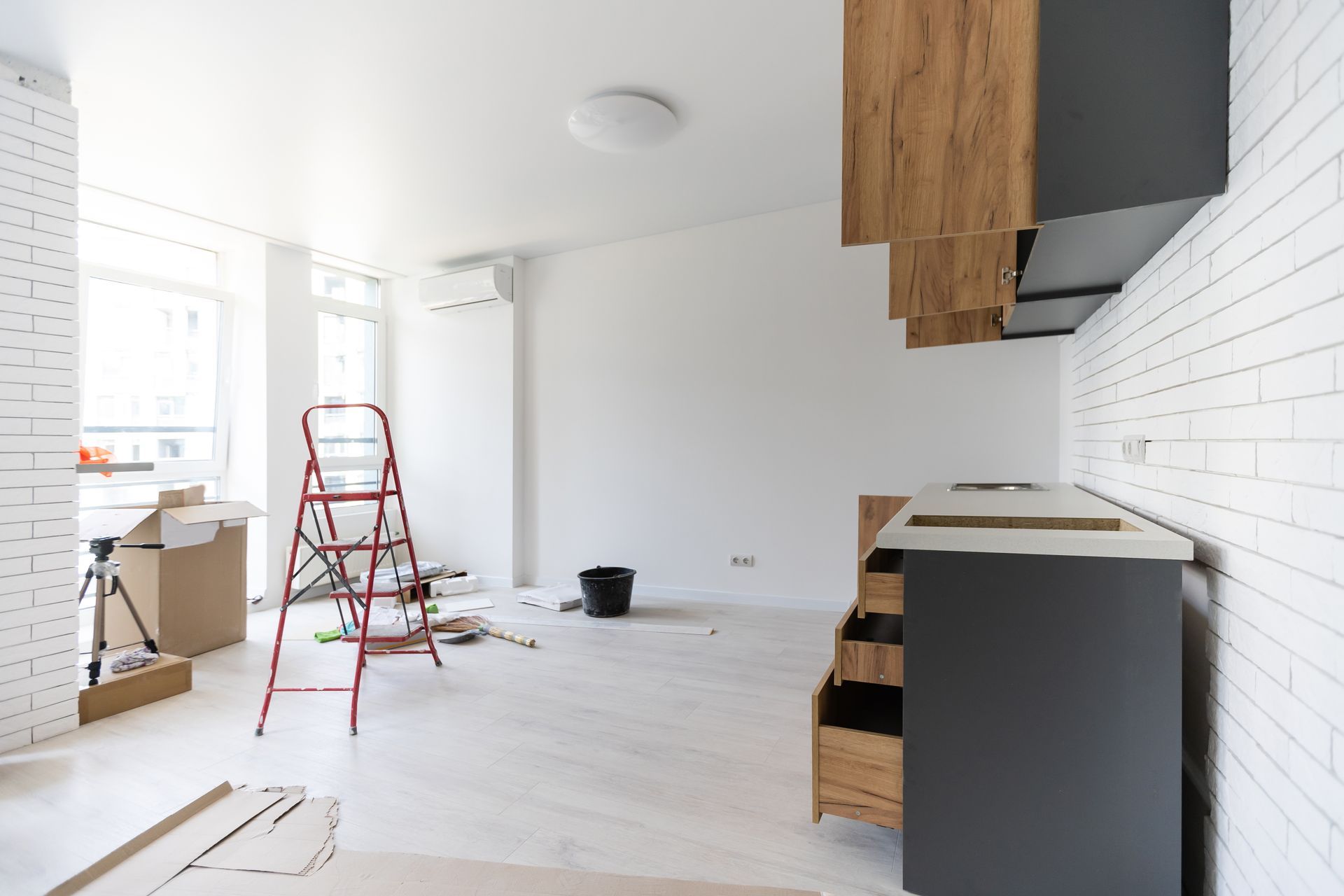 Interior with unfinished kitchen renovation; white walls, gray cabinets, exposed brick, ladder.