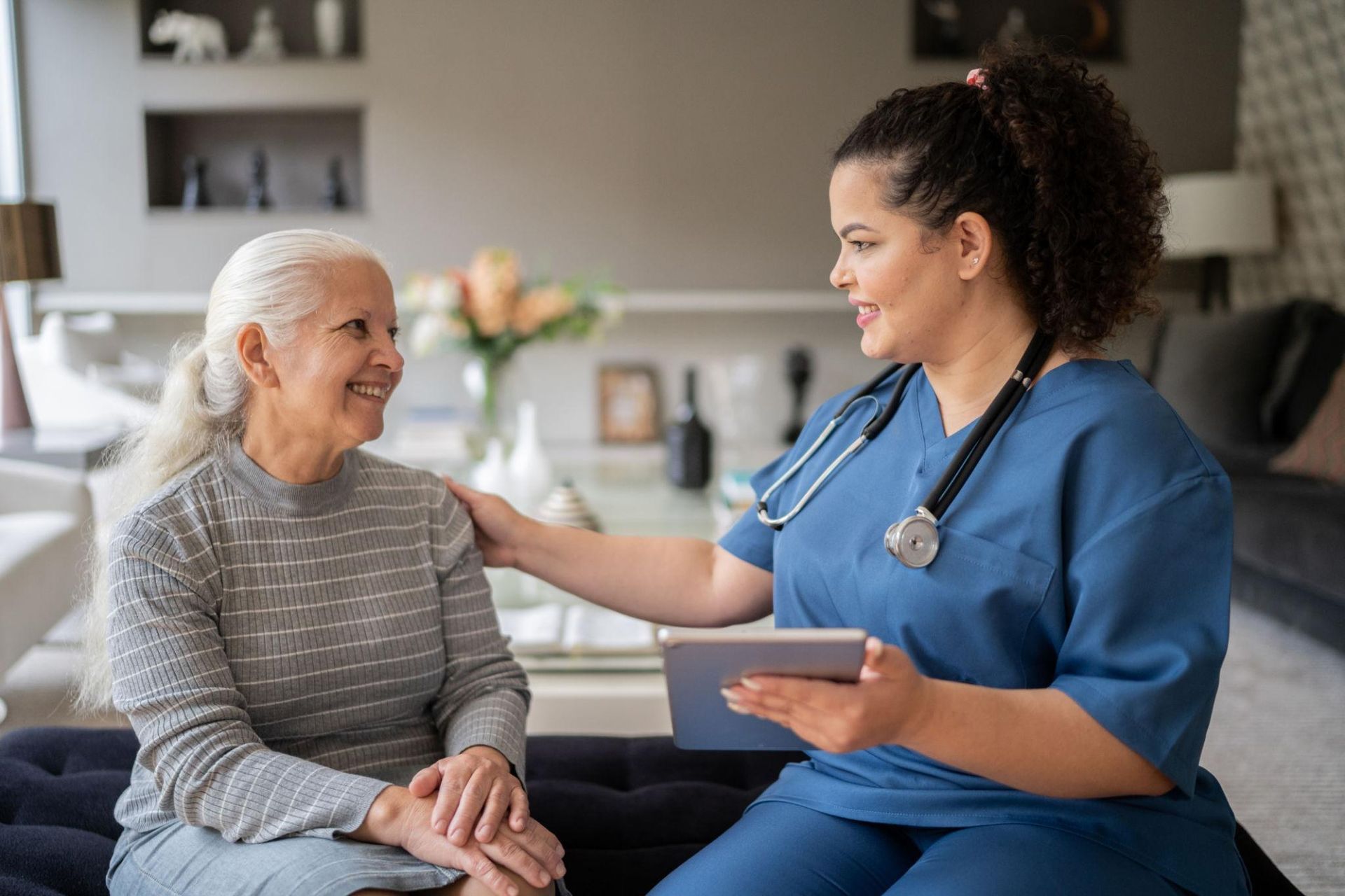 A healthcare worker smiles and points while speaking to an elderly person in a bright room.