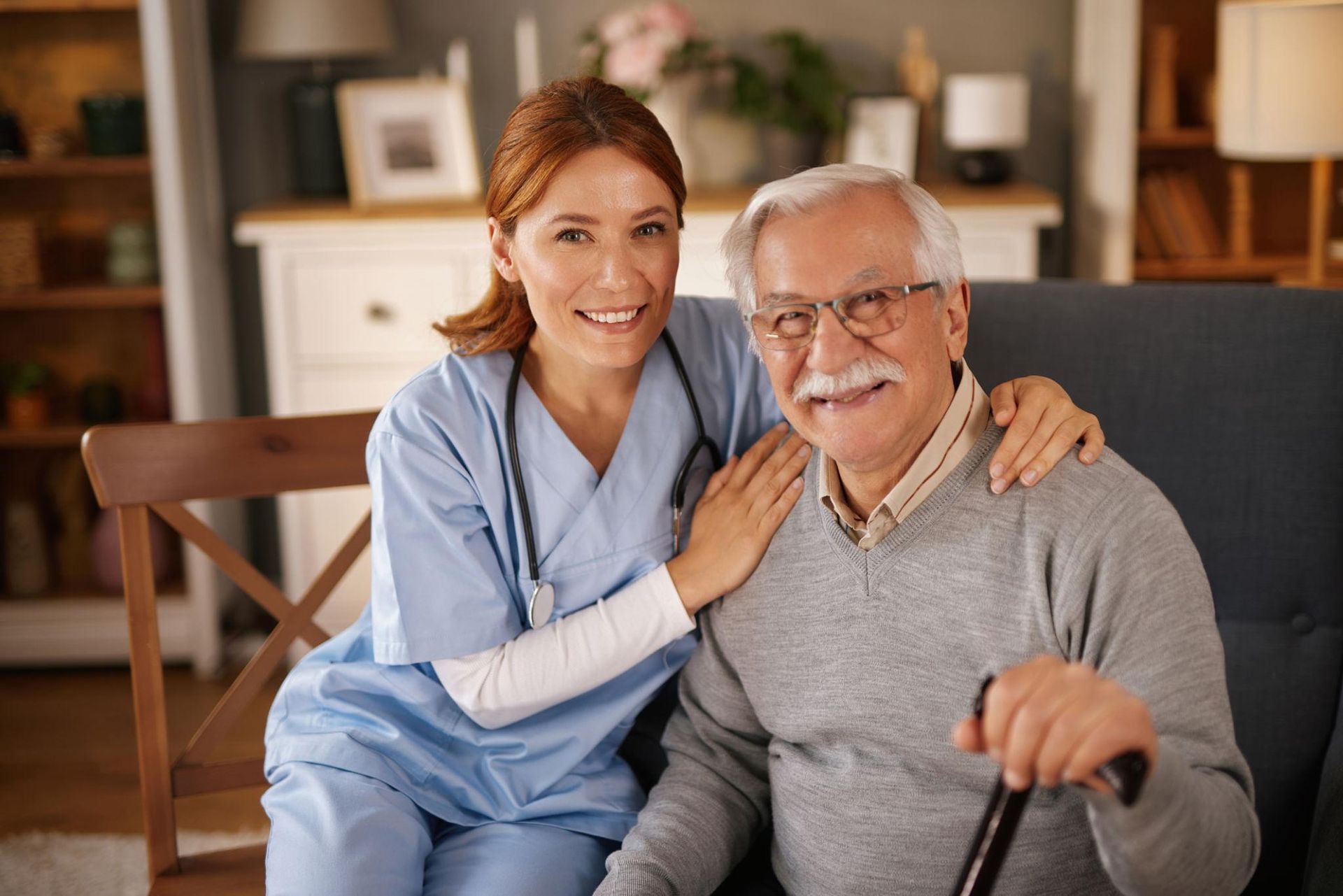 A healthcare worker smiles and points while speaking to an elderly person in a bright room.