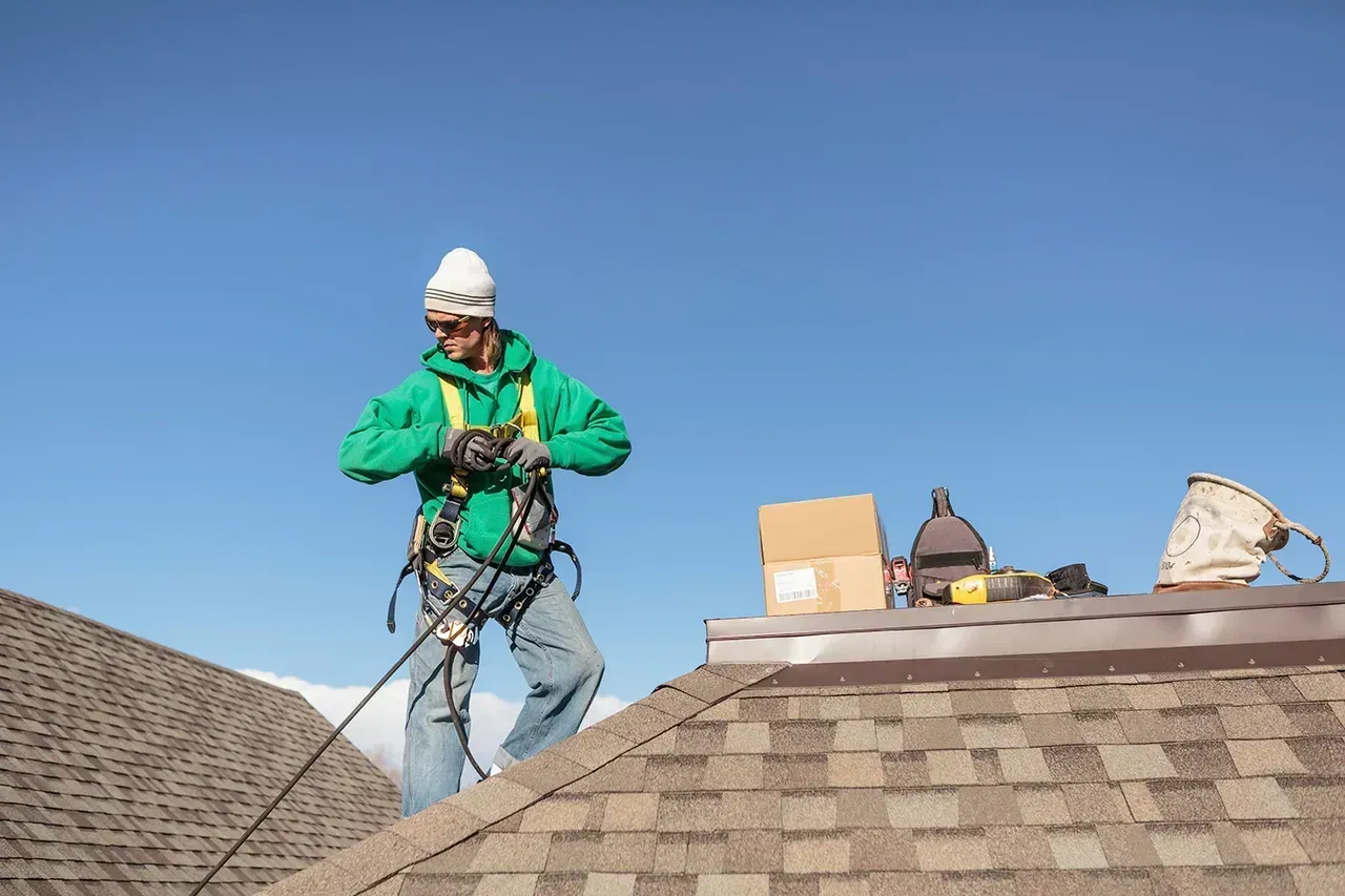 Roofer in green jacket and hard hat on shingled roof beside tools and materials under a clear blue sky