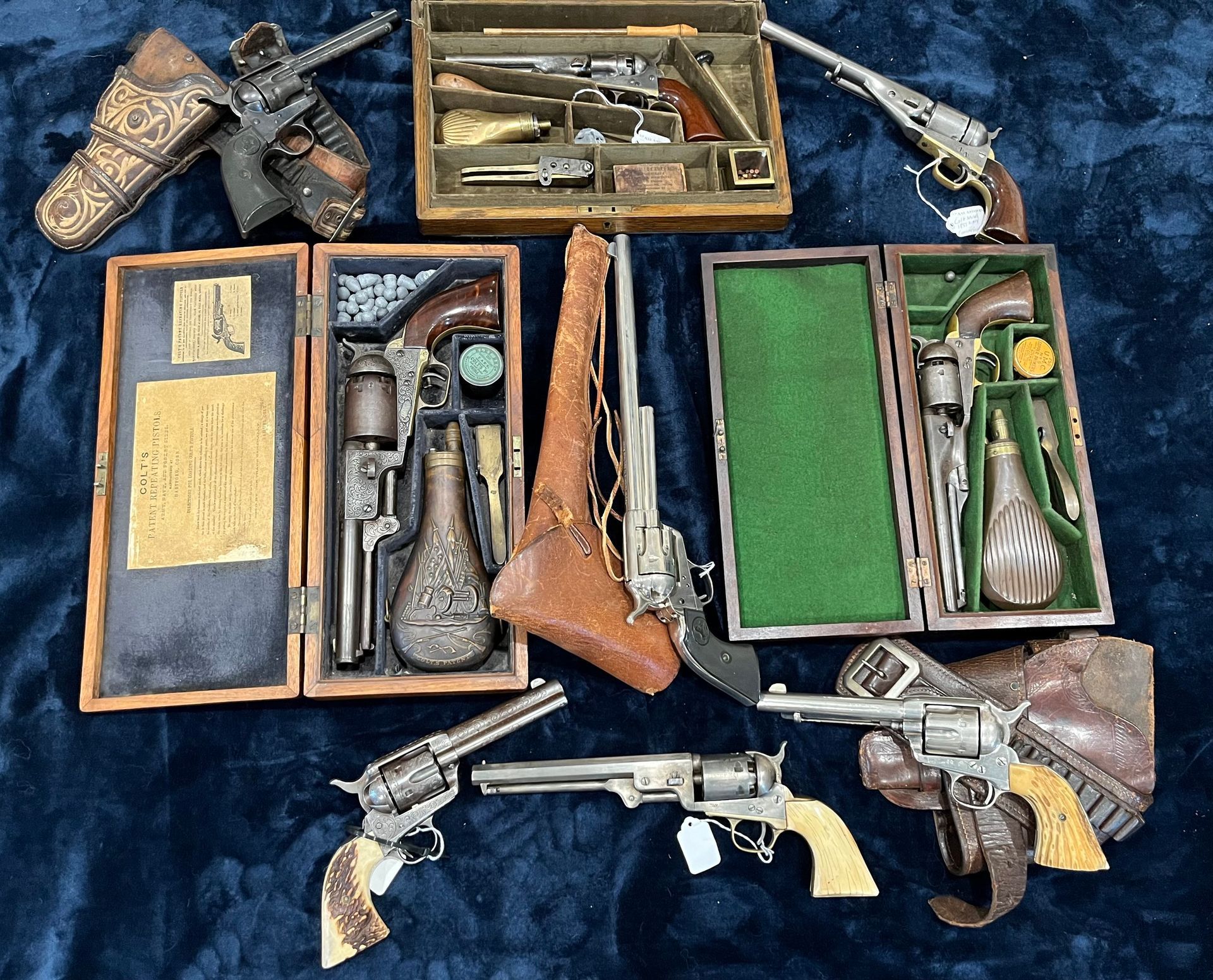 Various antique firearms, including revolvers and a long gun, displayed on a dark blue surface. Some are in wooden cases or holsters.