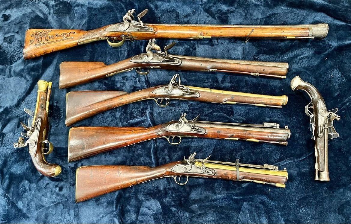 Several antique flintlock firearms, including rifles and pistols, displayed on a dark blue surface. Brown wood and silver metal are prominent.