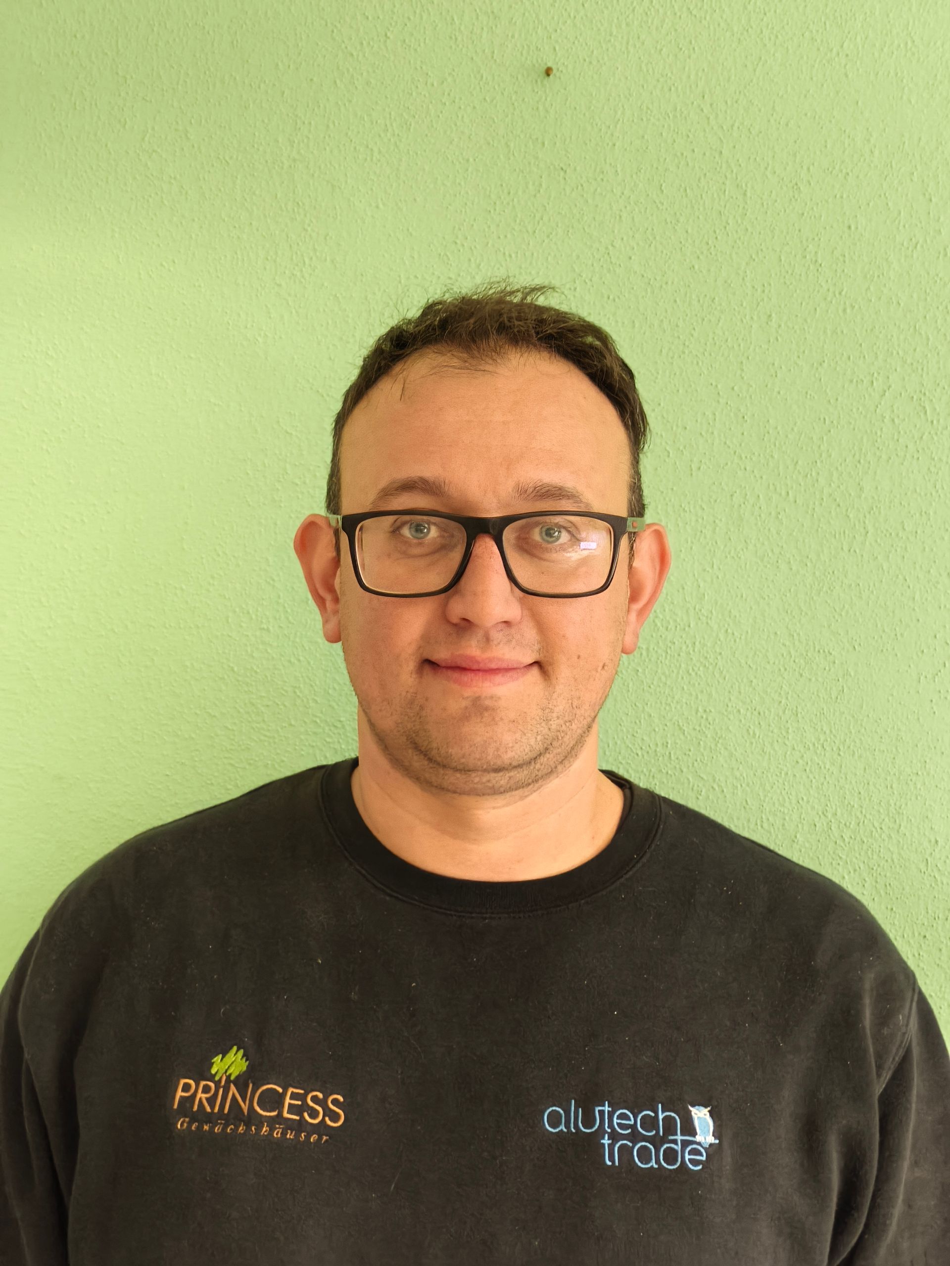 Frau mit Brille und langen Haaren in einem schwarzen Hemd posiert vor einer grünen Wand.
