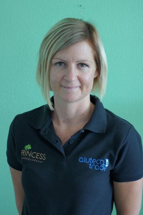 Eine Frau mit blonden Haaren, die ein schwarzes Poloshirt trägt, steht vor einer türkisfarbenen Wand.