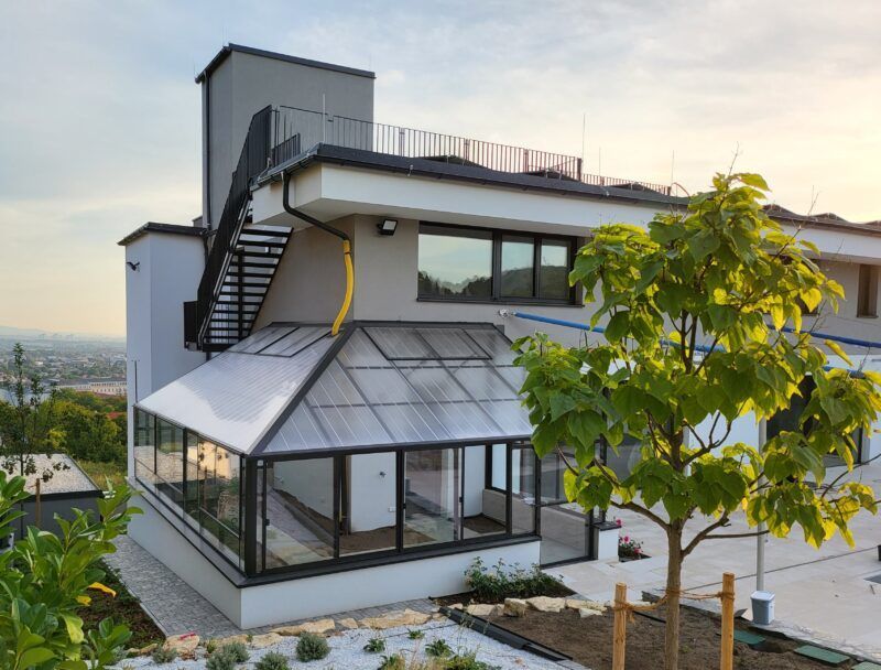 Modernes Haus mit gläsernem Wintergarten, Außentreppe und Dachterrasse vor dem Hintergrund eines Sonnenuntergangs.