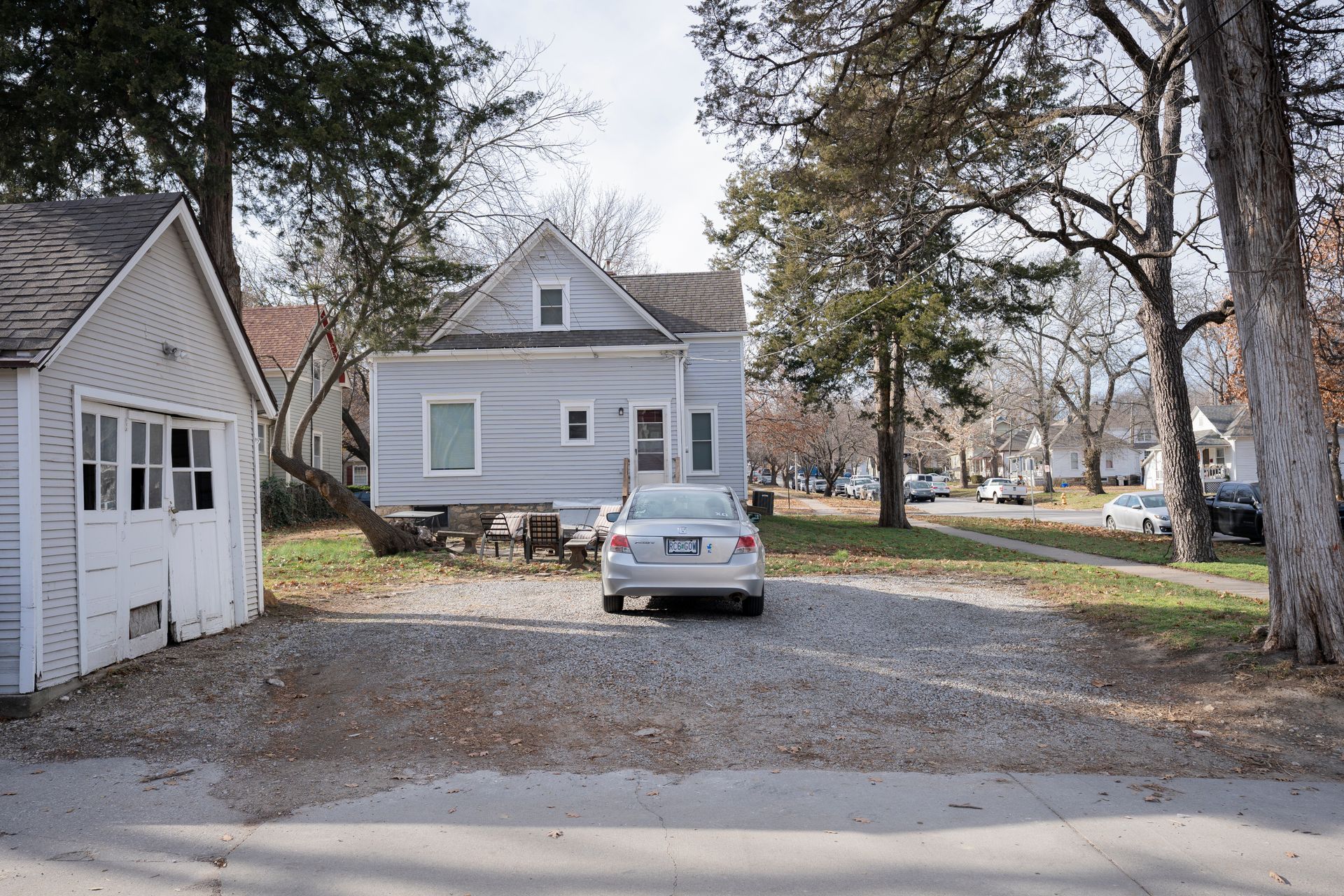 A small, light gray house with a car parked in front of it, near a tree-lined street.