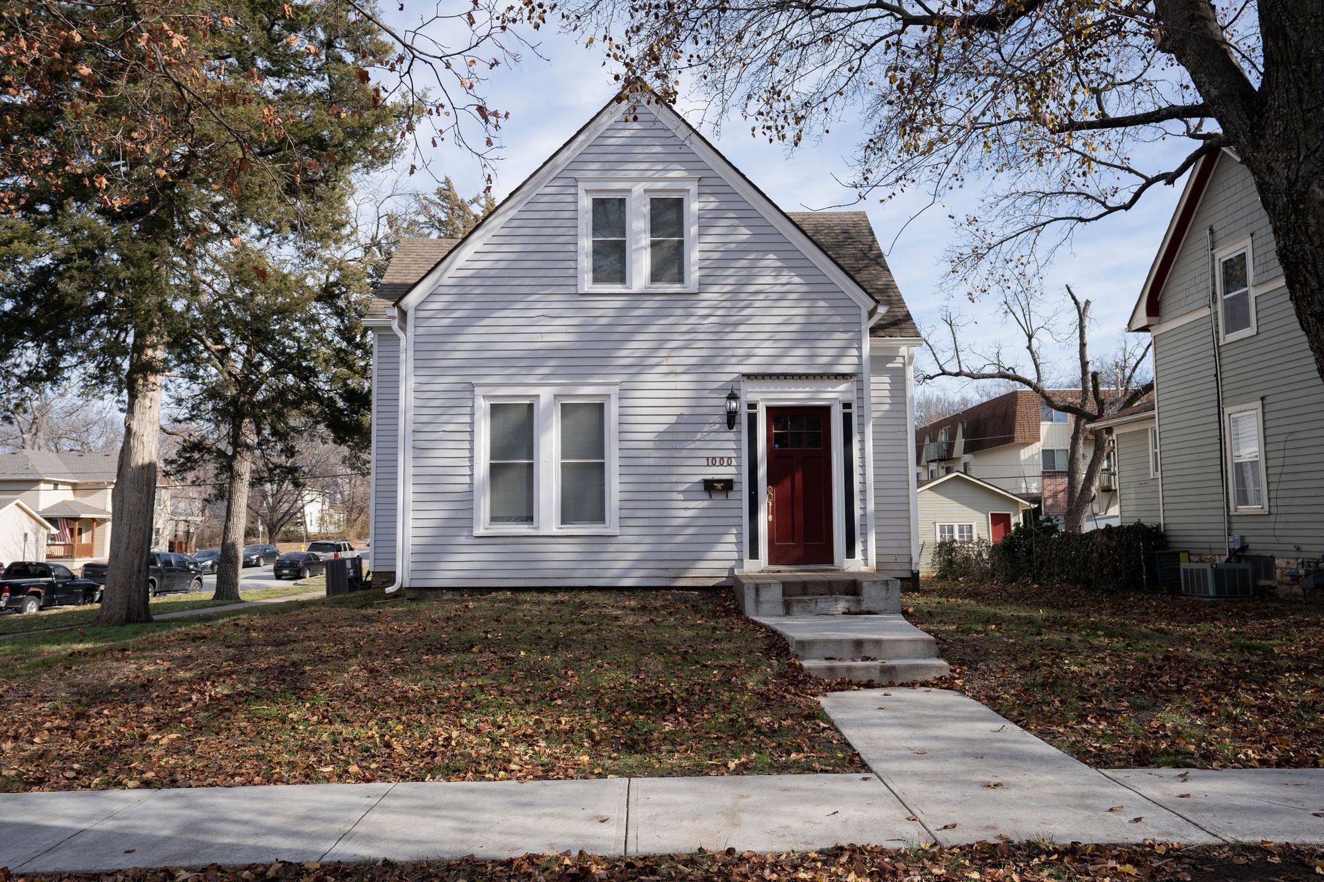 Light blue house with red door, sidewalk, trees in the yard, and another house visible to the right.