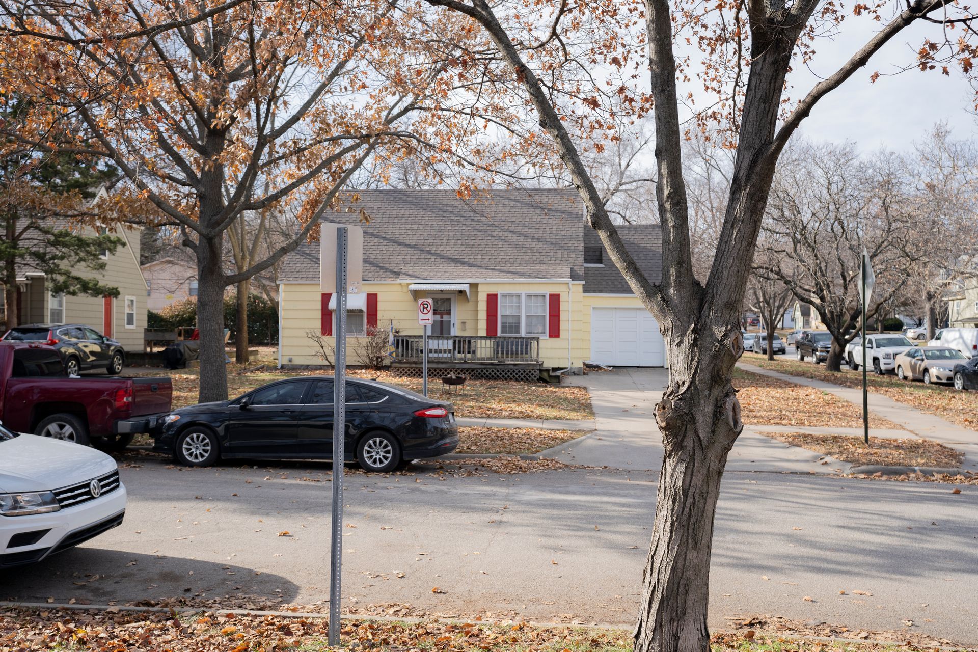 Yellow house with red trim, trees, parked cars, and fallen leaves on street.