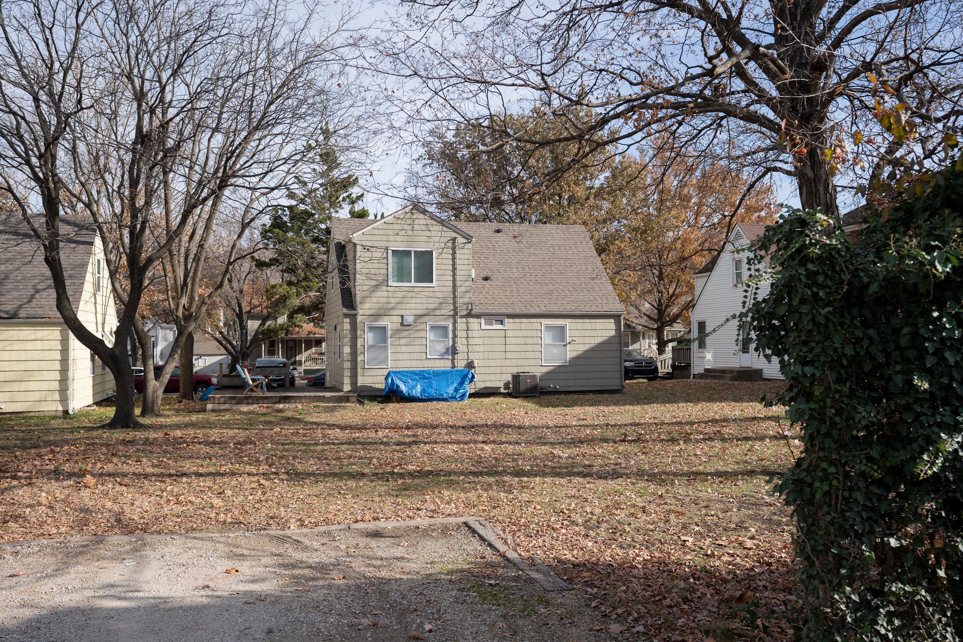 A two-story house with a beige exterior and brown roof, surrounded by bare trees and autumn leaves.