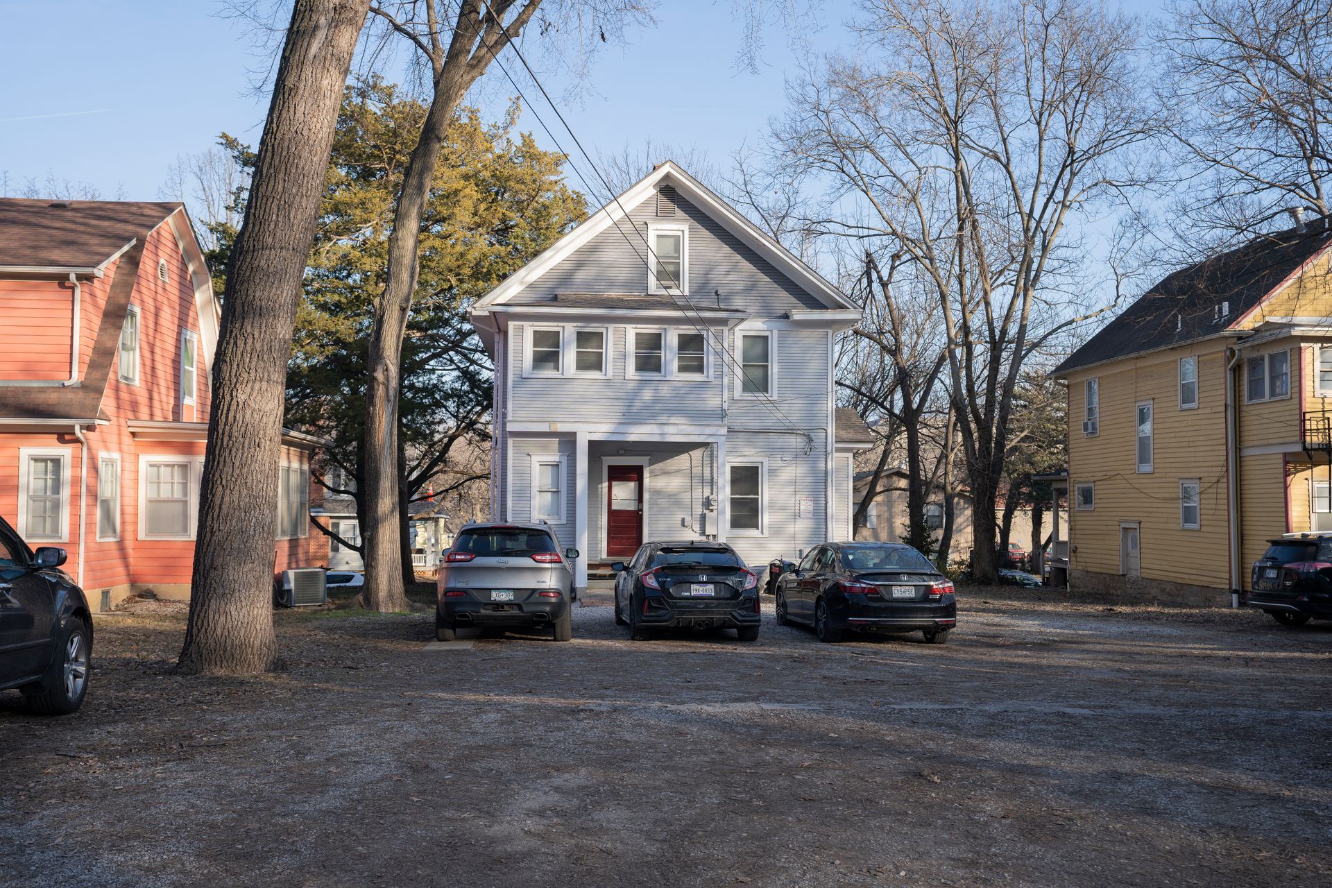 A light blue two-story house with a red door, surrounded by parked cars, on a gravel lot.