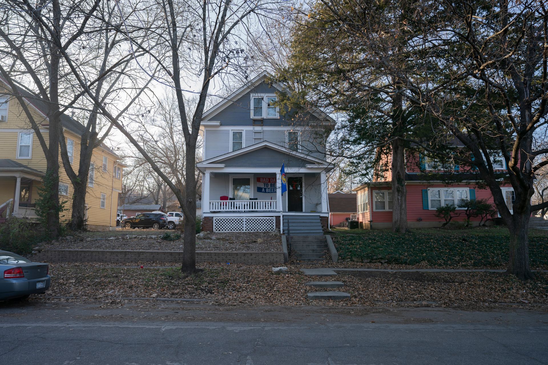 Two-story light blue house with porch, trees, and cars on a street.