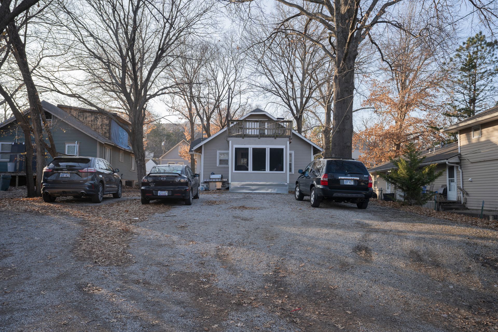 Cars parked in front of a gray house with bare trees in a gravel driveway, possibly autumn.