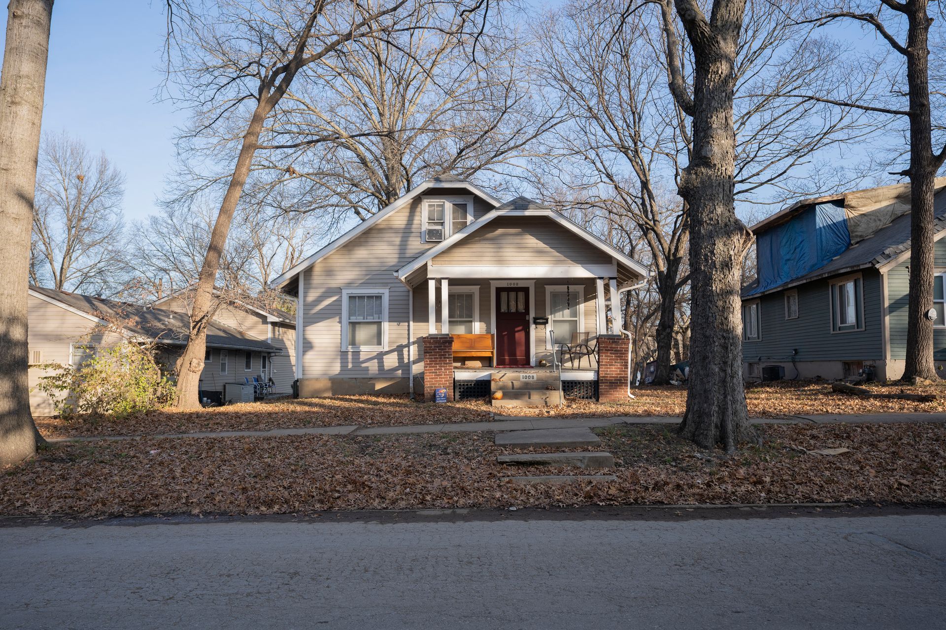 Small house with a porch and red door, flanked by trees, on a residential street.