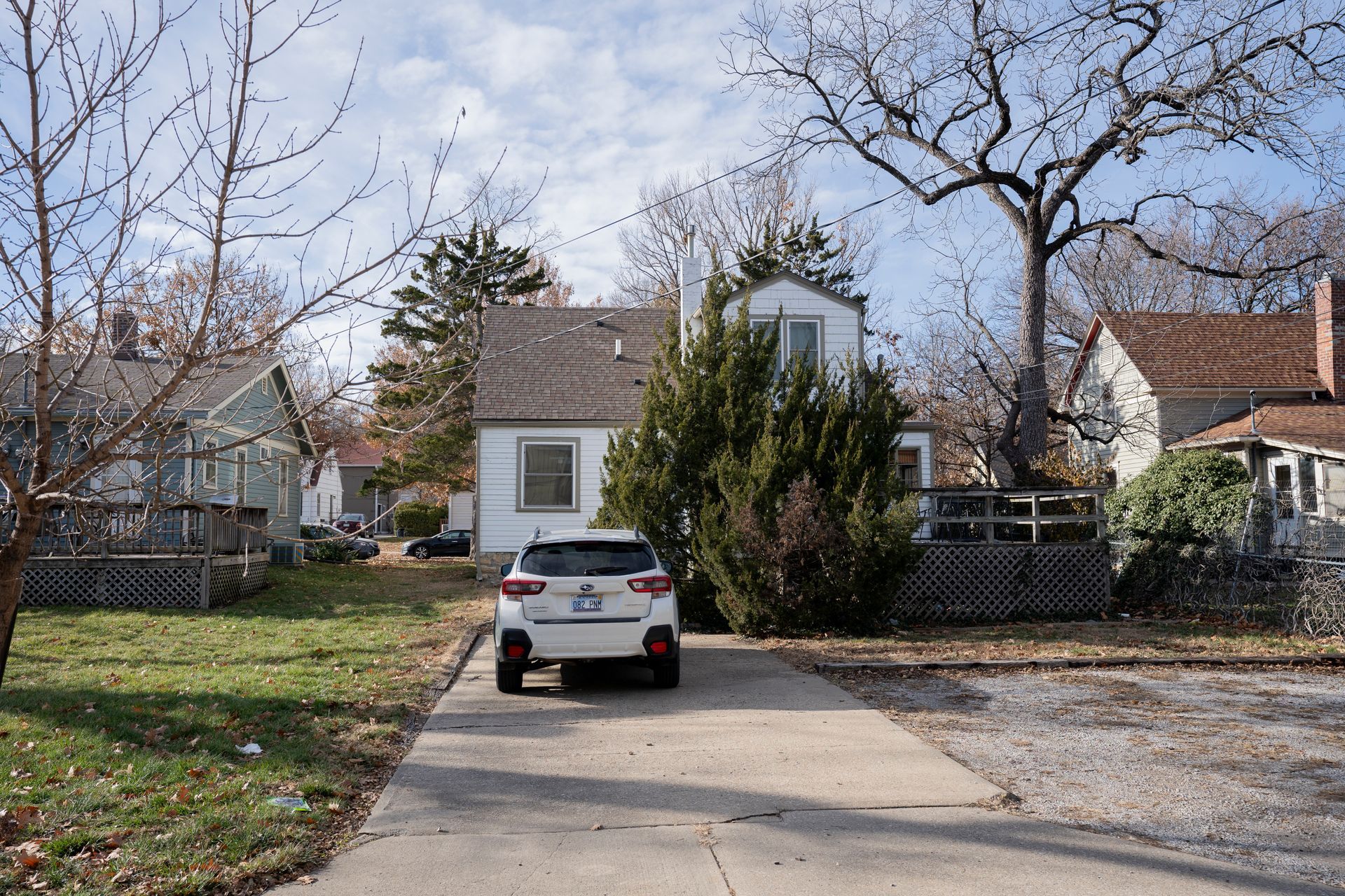 White car parked in front of a white house on a concrete driveway, surrounded by other houses and bare trees.