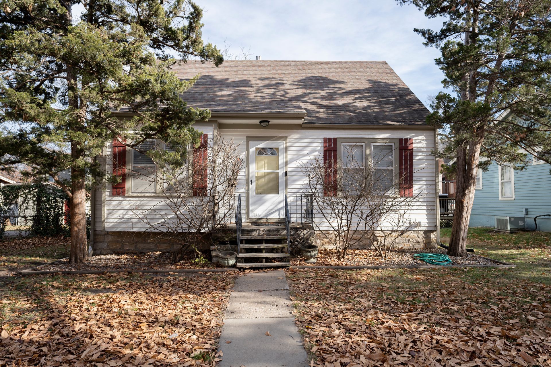 Small white house with red shutters and a brown roof, trees on either side.