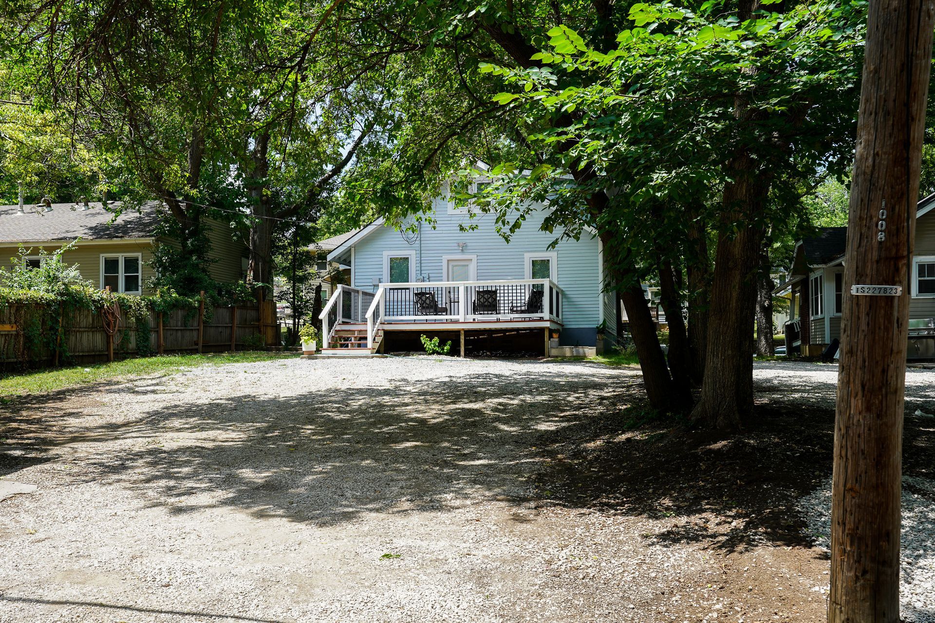 Gravel parking area in front of a light blue house with a porch. Trees partially obscure the view.