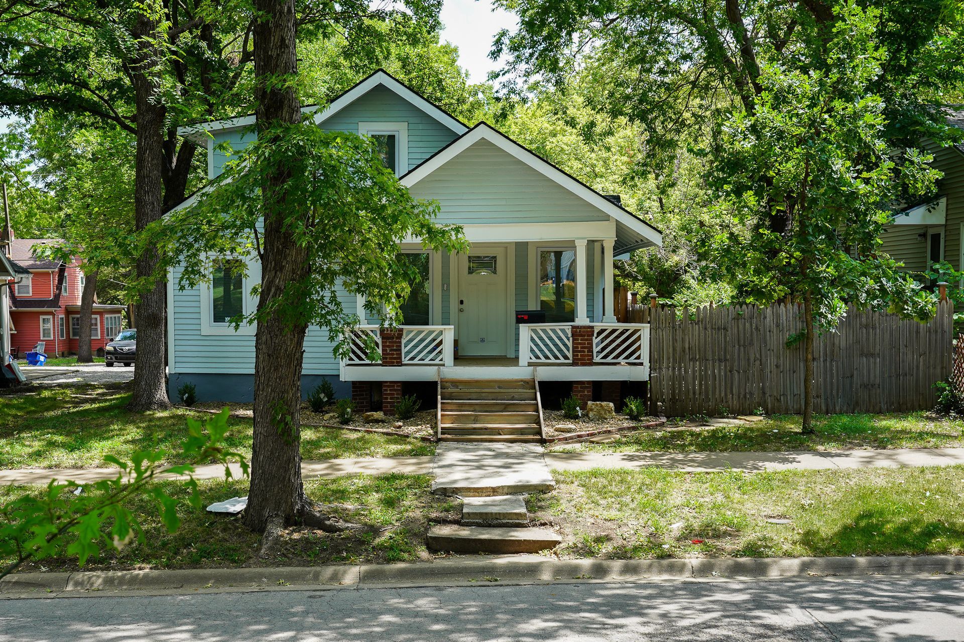 Light blue house with porch, steps, and trees. Green grass and a sidewalk are in front.