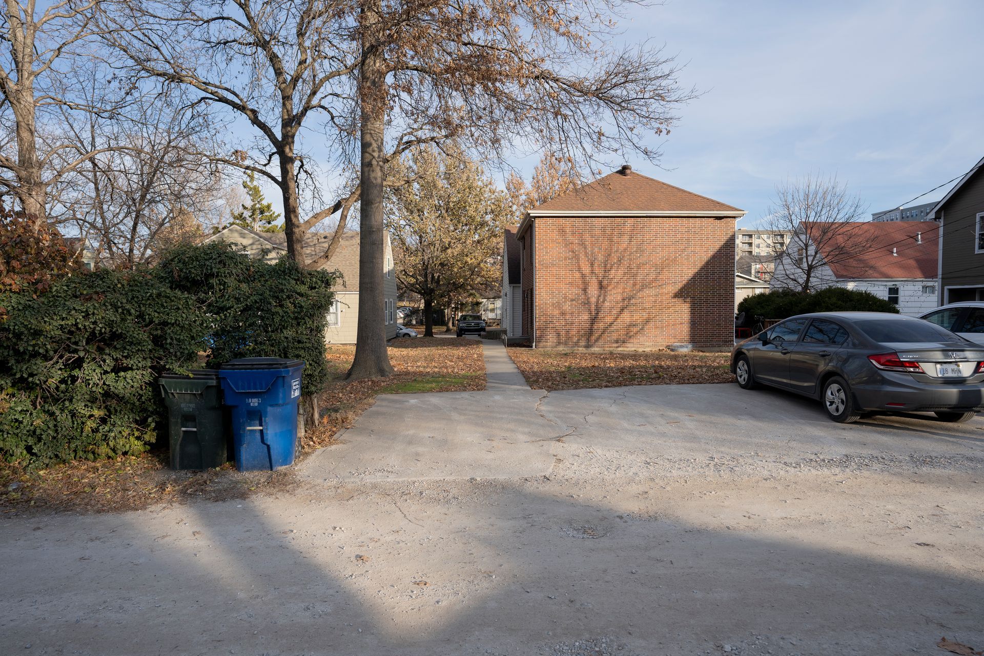 Gravel driveway with a parked car, trash bins, and a brick building in the background under a sunny sky.