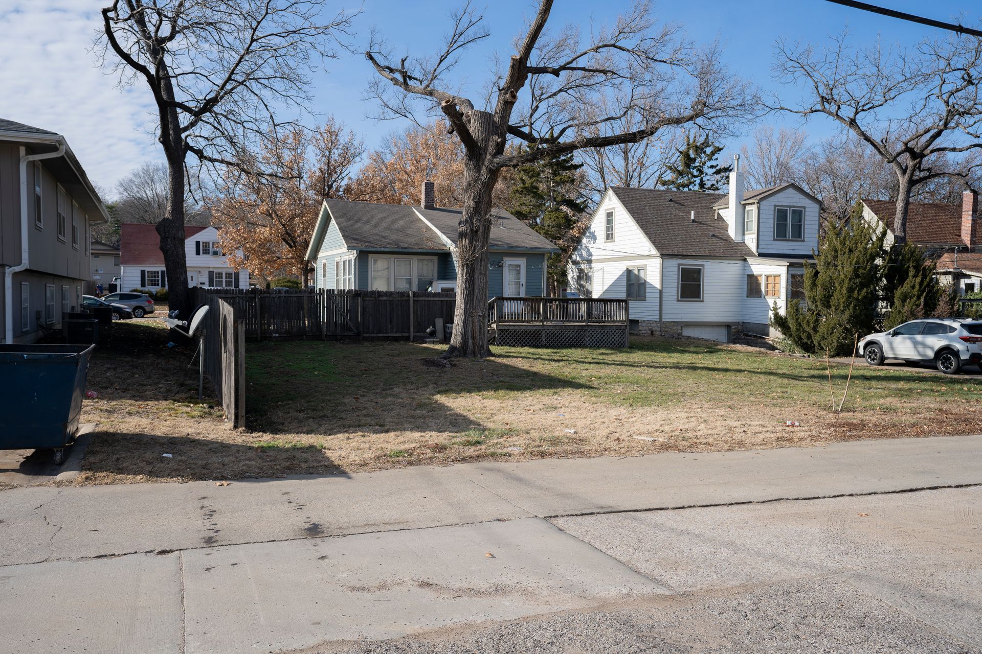 Houses along a street on a sunny day; trees with bare branches, cars parked along the side.