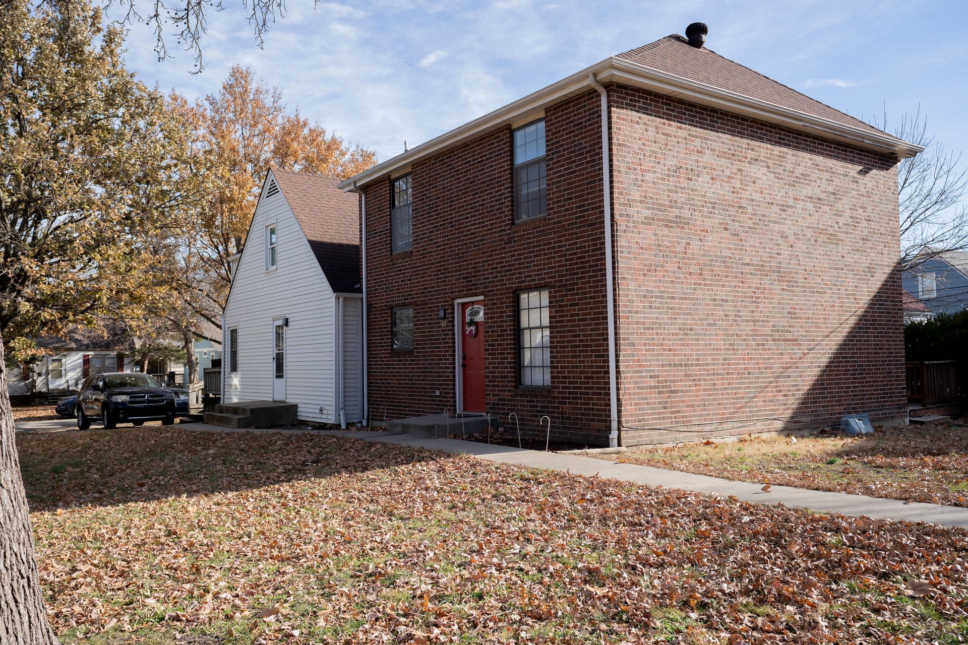 Two-story brick house with a red door next to a smaller white house on a fall day.
