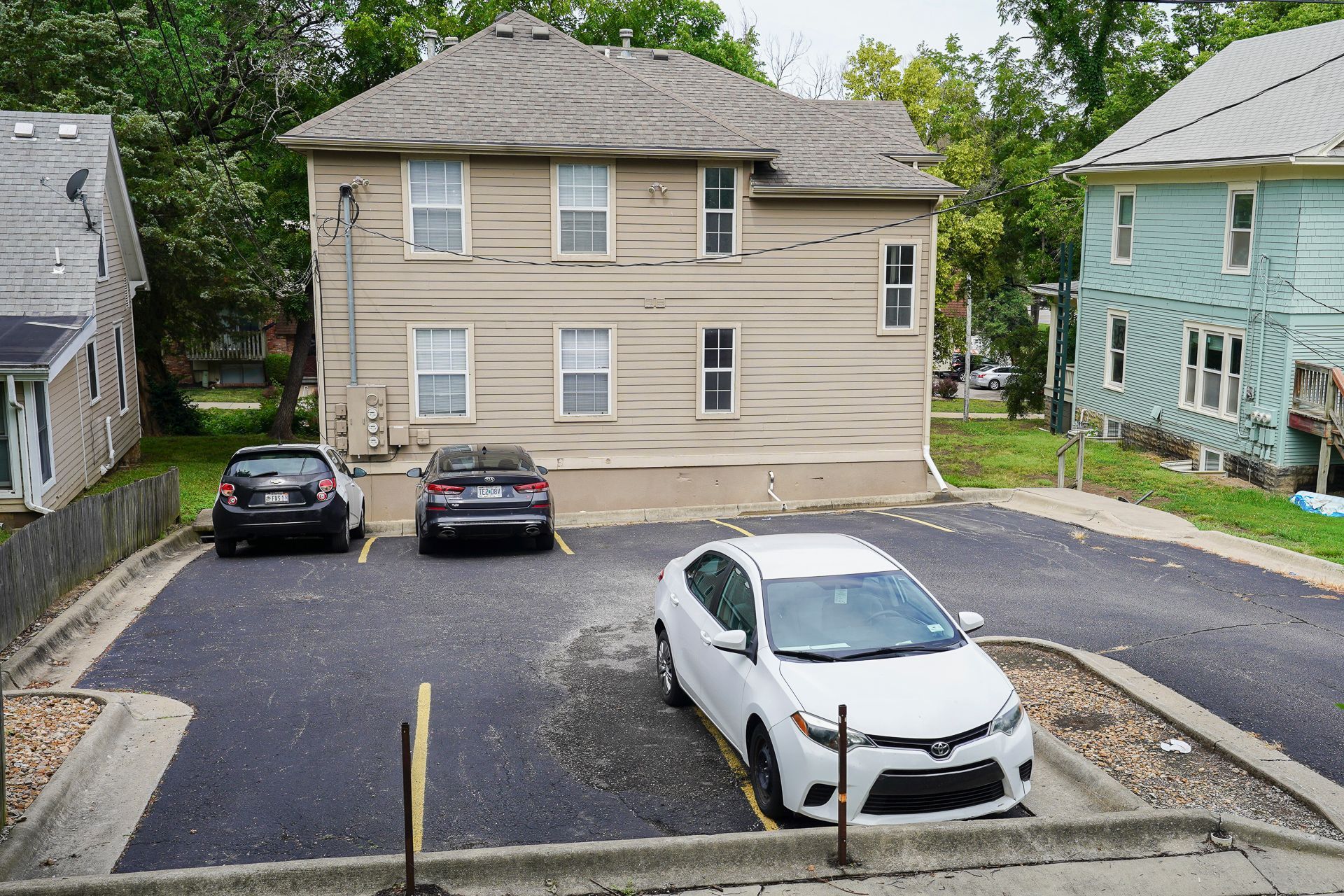 Three-story beige building with a parking lot in front, cars parked. Green trees in the background.