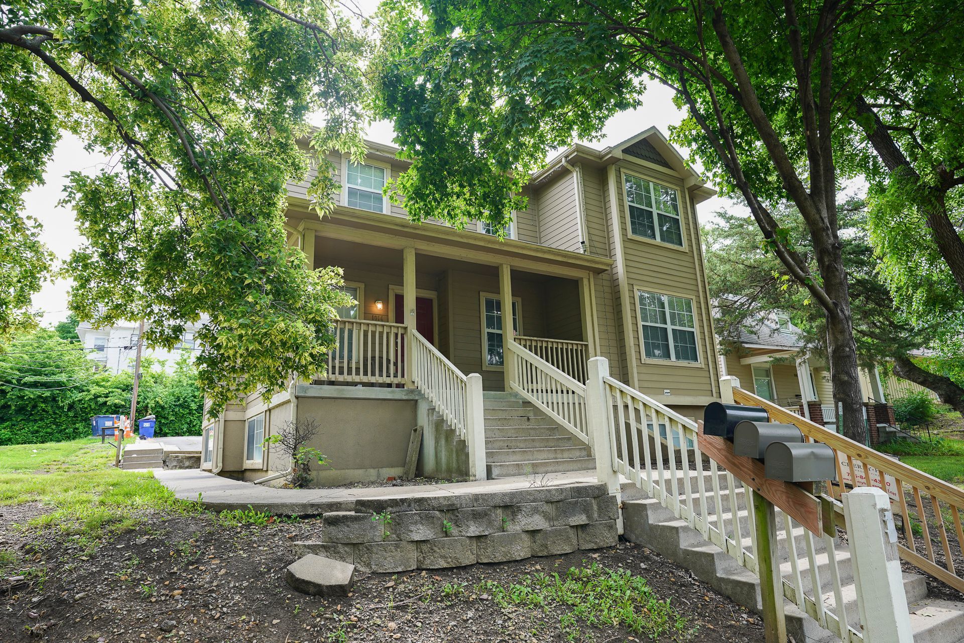 Two-story beige house with porch, stairs, and mailboxes. Overgrown with trees and green grass.