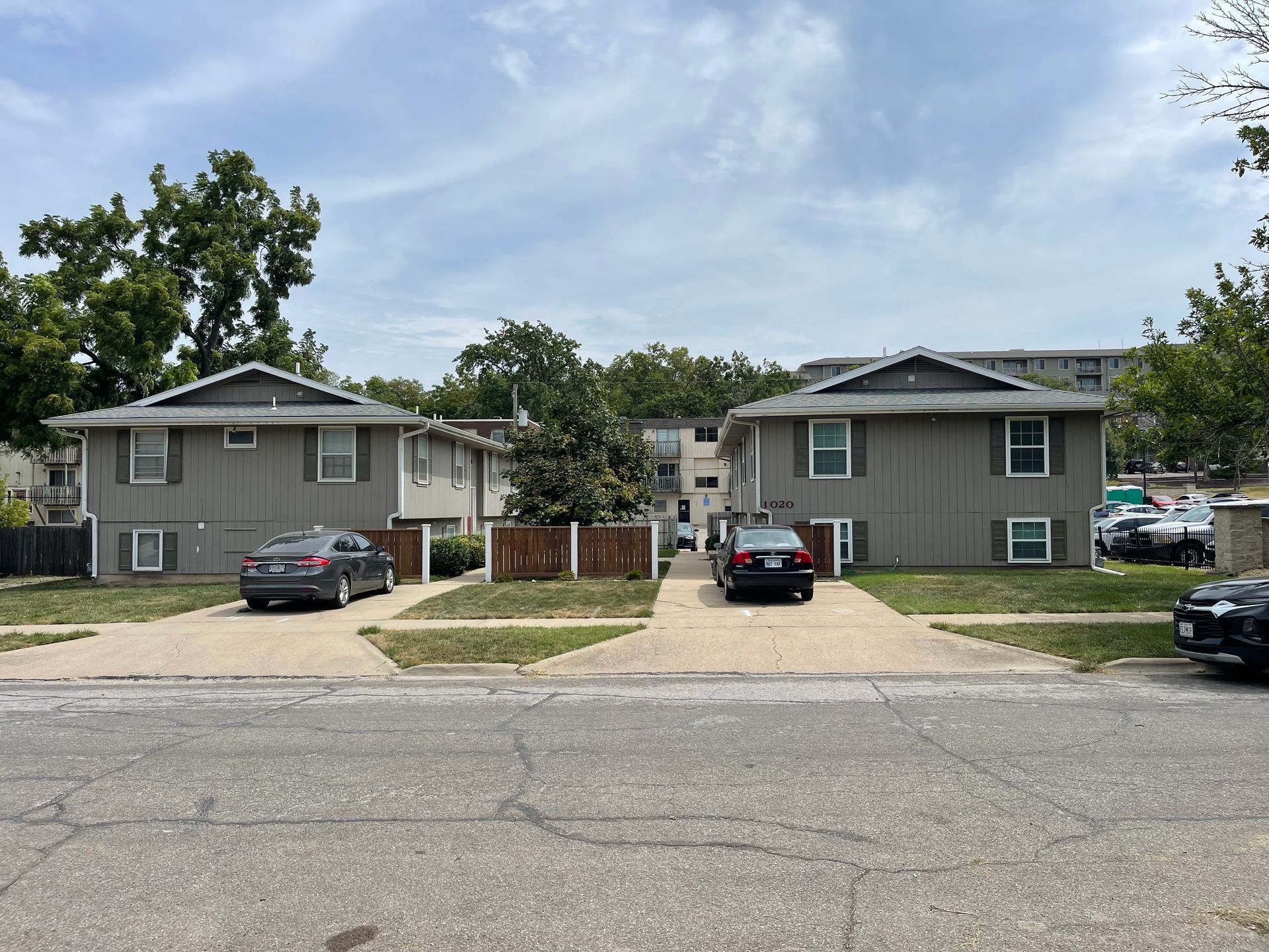 Two gray multi-unit houses with cars parked in front on a sunny day.