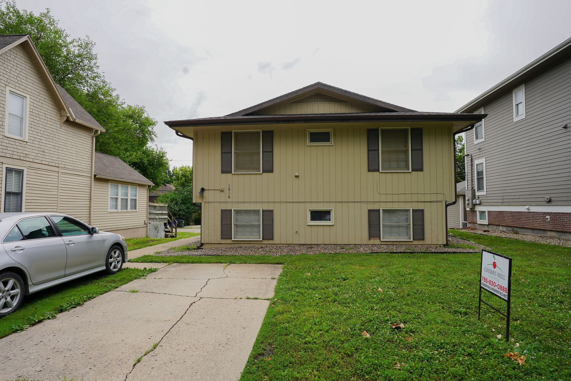 Two-story tan house with brown shutters and a driveway, parked silver car, and a for sale sign.