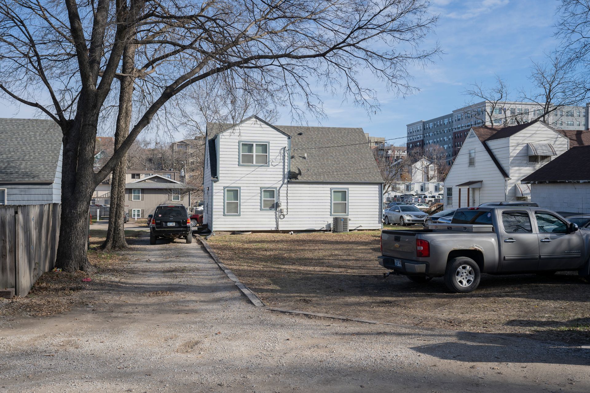 A gravel driveway with cars parked, leading to a white house in a residential neighborhood on a sunny day.