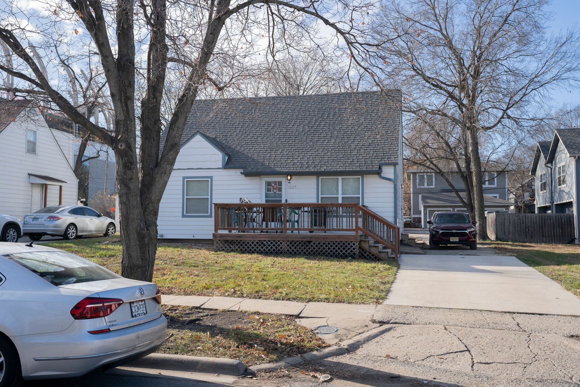 White house with a wooden deck and driveway on a residential street. Cars and trees are also visible.
