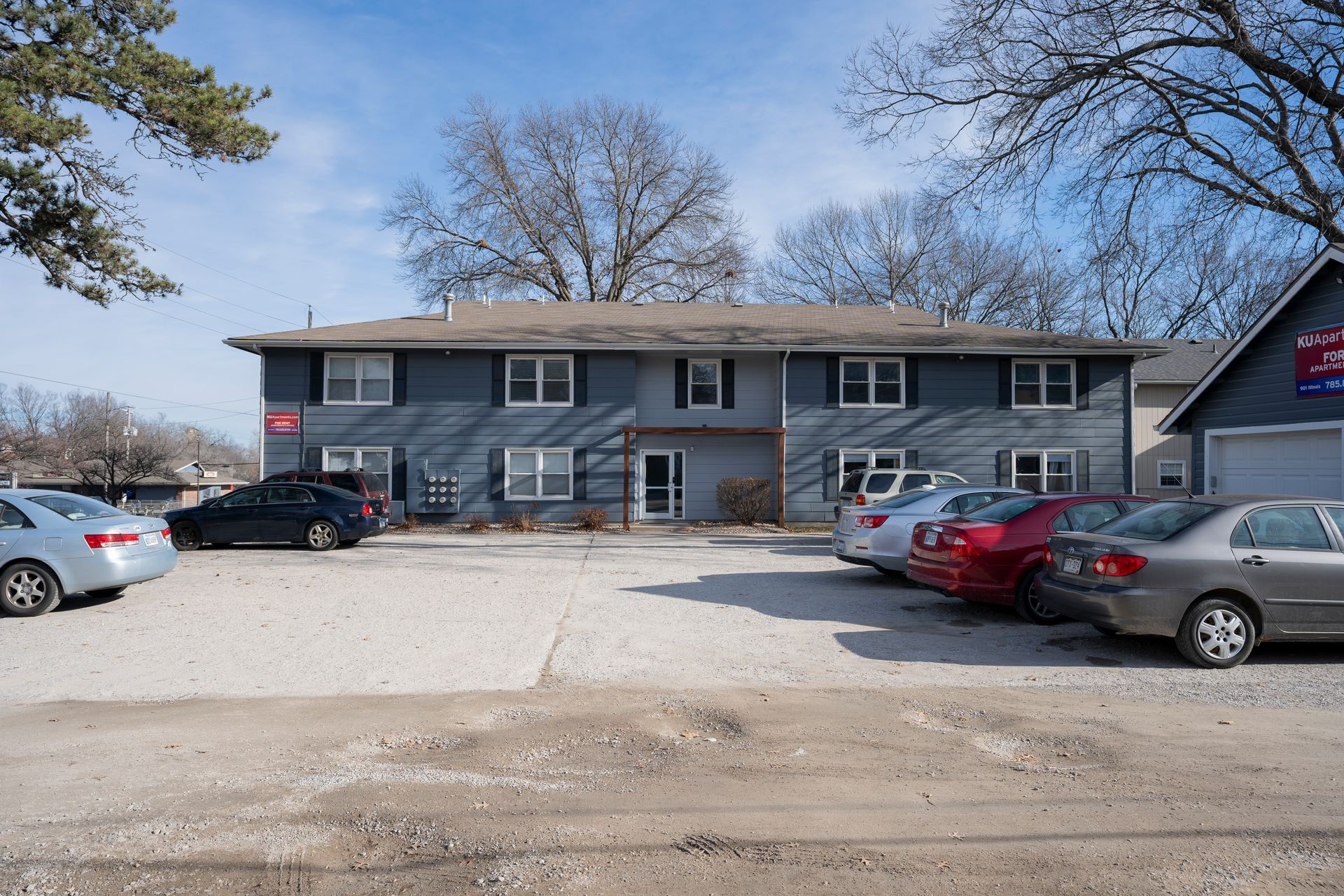Apartment building with several cars parked in front on a gravel lot on a sunny day.