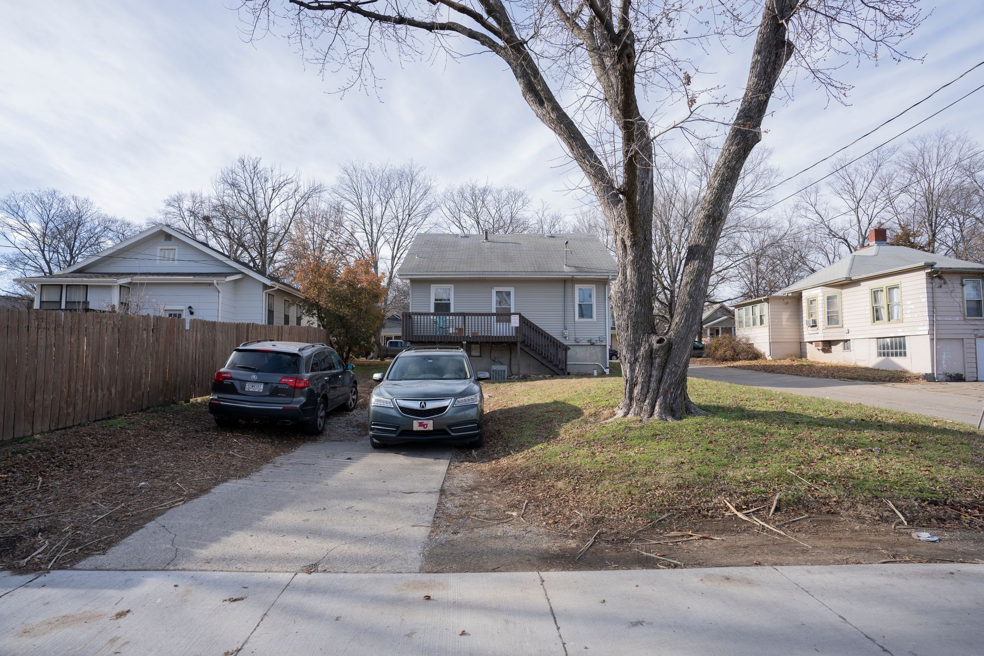Cars parked in front of a small house with a wooden deck, driveway, and street, trees in the background.