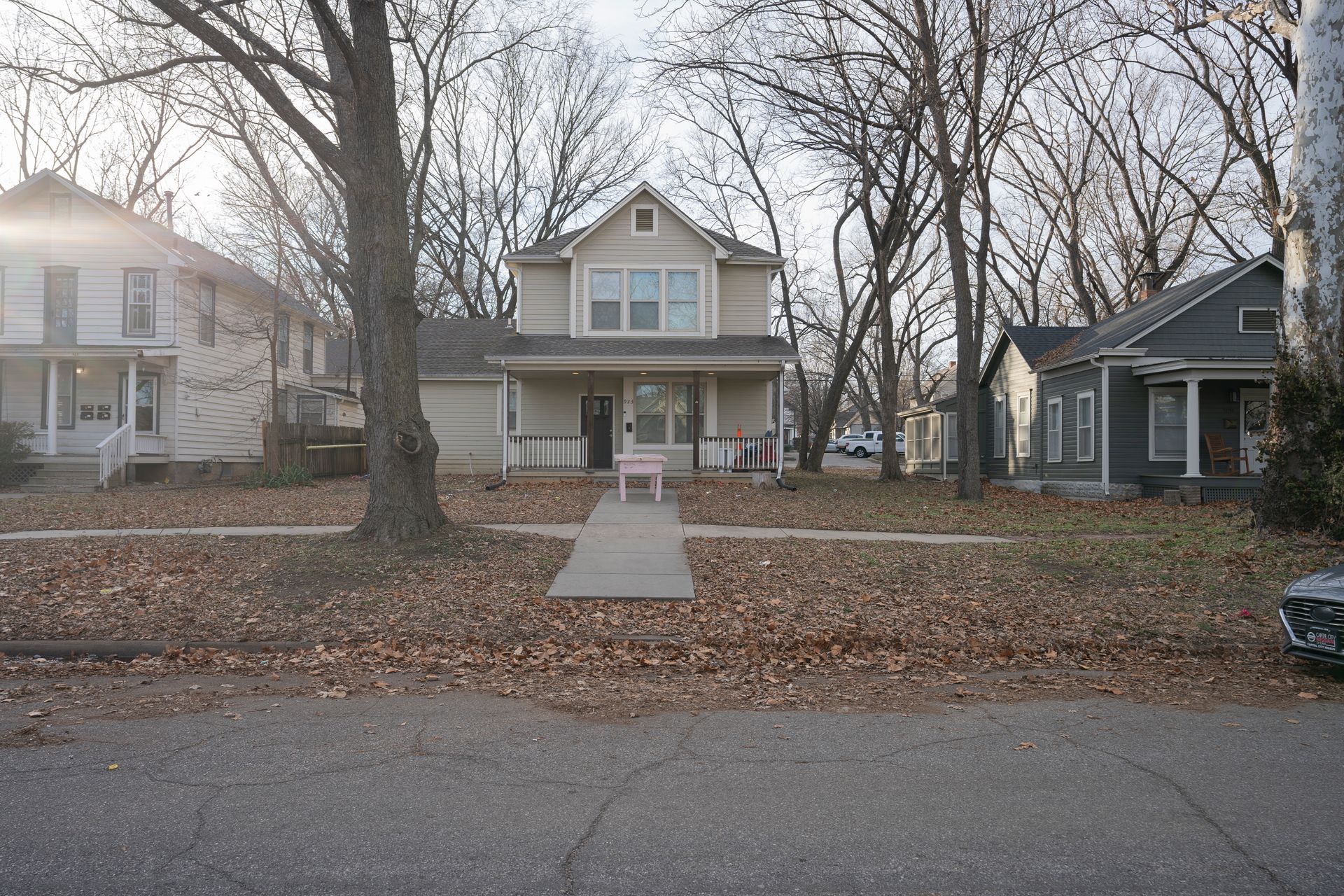 Houses on a street with bare trees and fallen leaves; a pink chair sits on a porch.