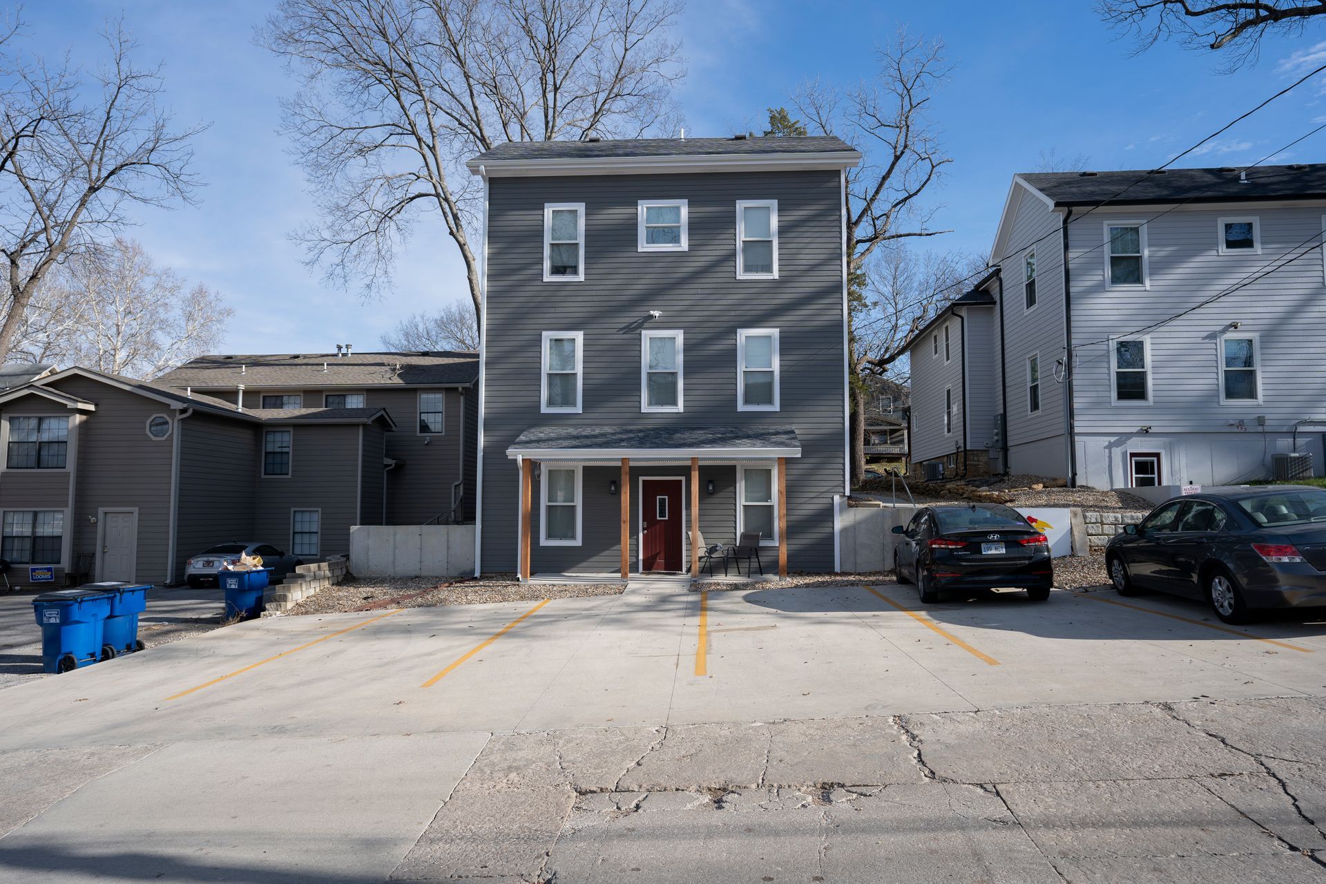 Gray apartment building with a parking lot in front. Blue trash bins on the left. Cars parked on the right.