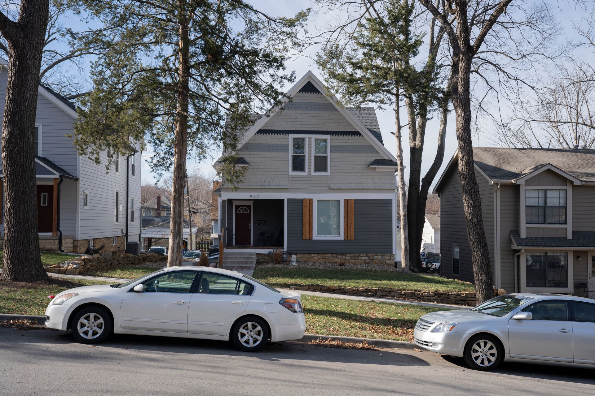 A two-story house with gray siding and a peaked roof, trees, and parked cars on a street.