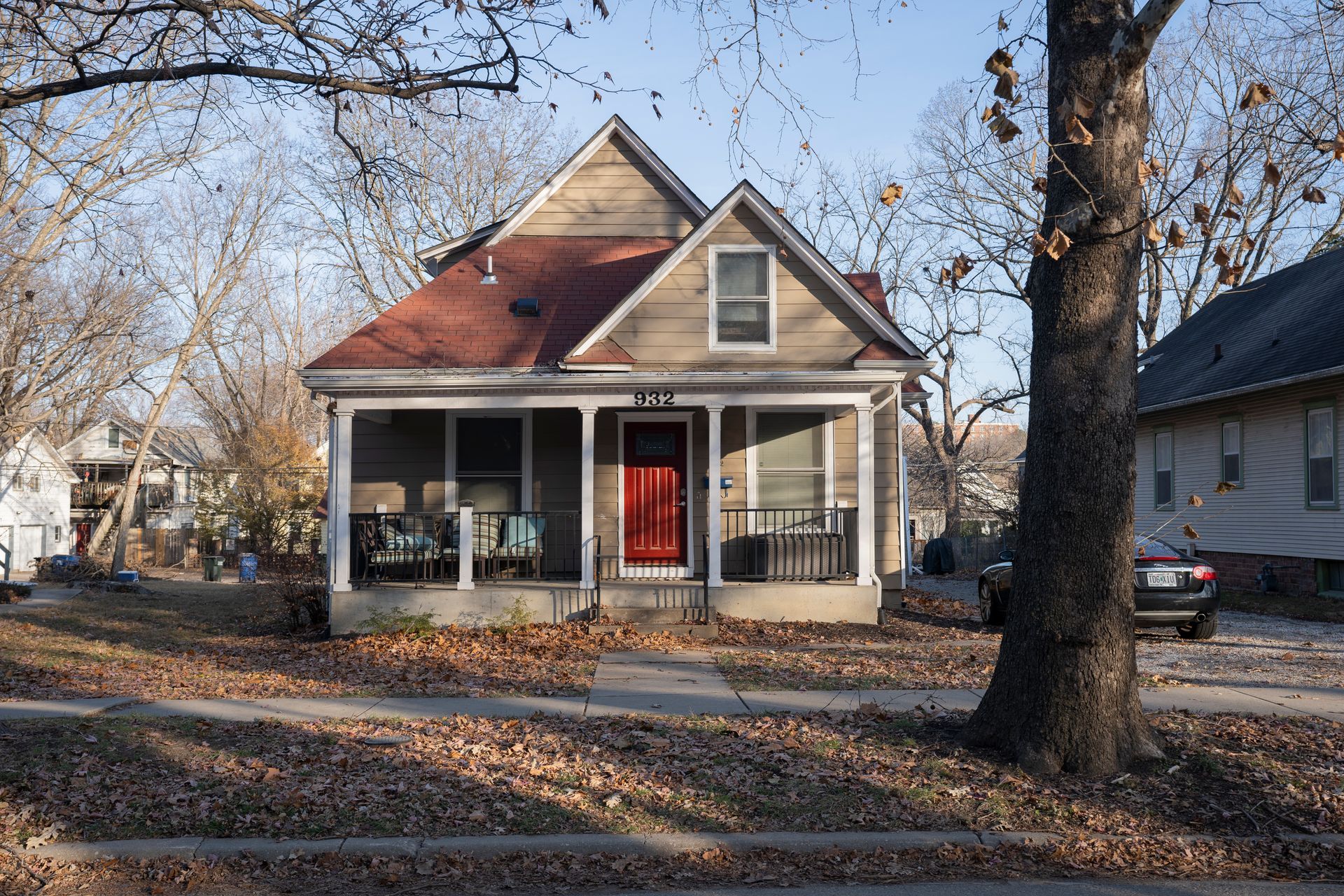 Small beige house with a red door and porch, fall leaves on the ground, trees.