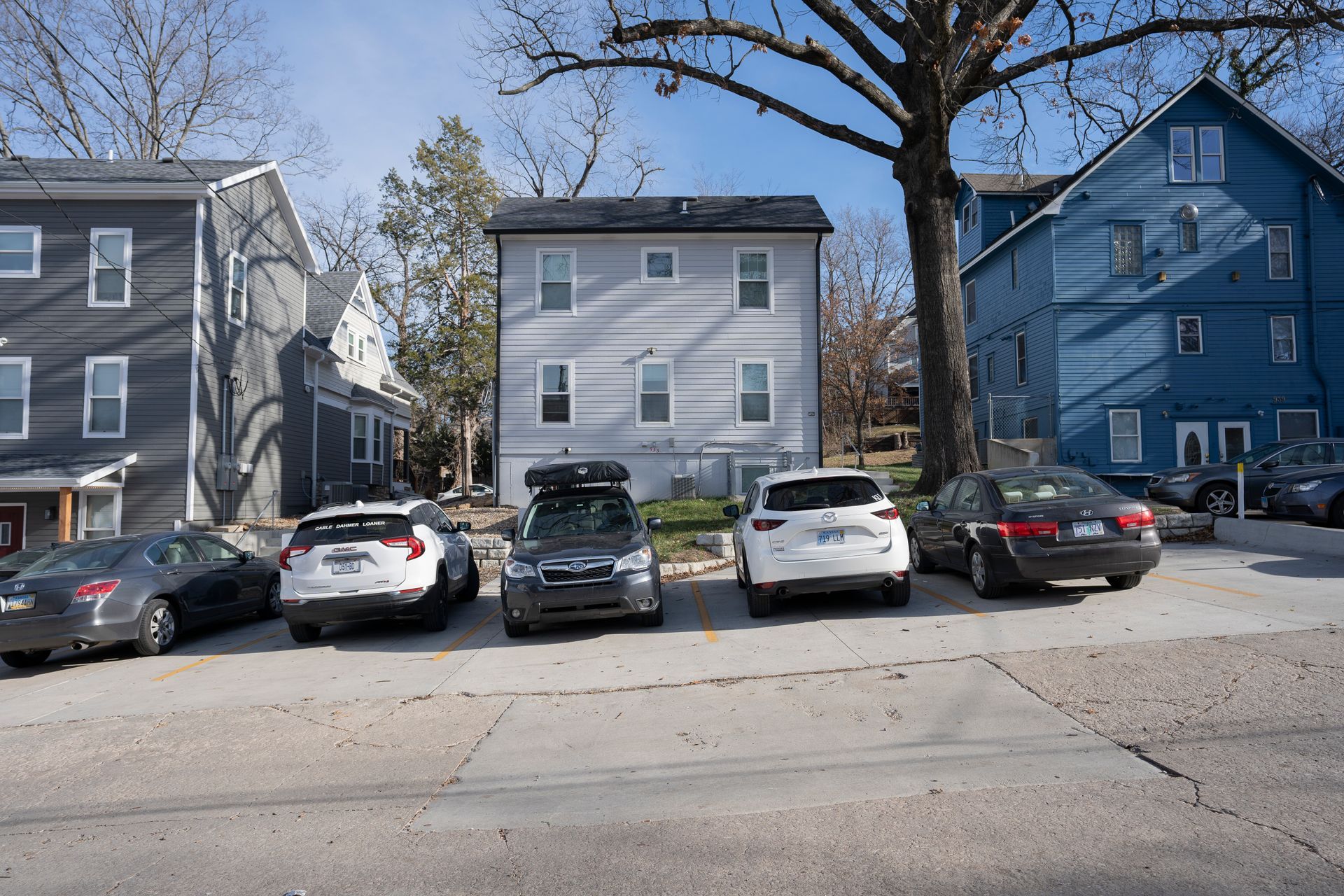 Cars parked in front of light grey and blue houses. A sunny day, bare trees.