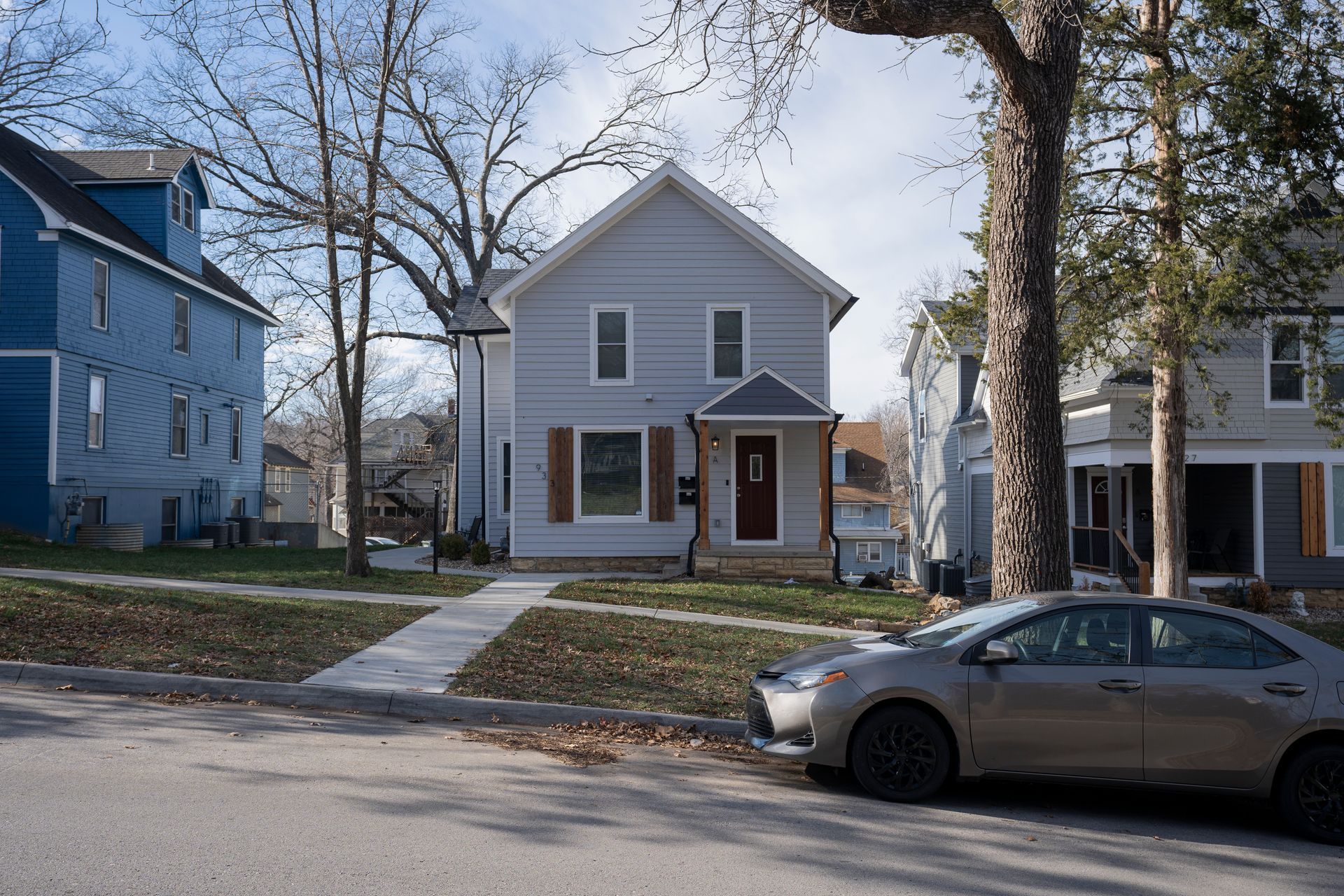 Light blue house with a dark red door, brown shutters, and a parked car on the street.