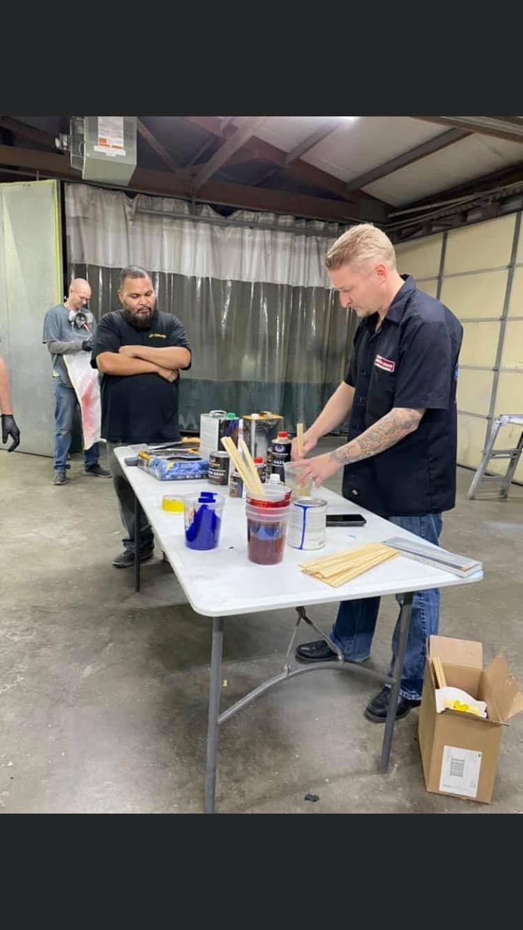 Two men at a table, working on items. One man has tattoos and examines something, the other watches. Workshop setting.