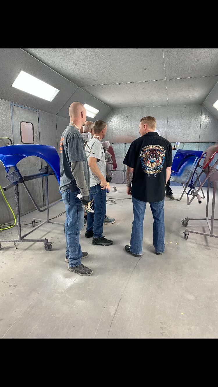 Three men in a body shop, talking near car parts. Blue and red car pieces are visible.