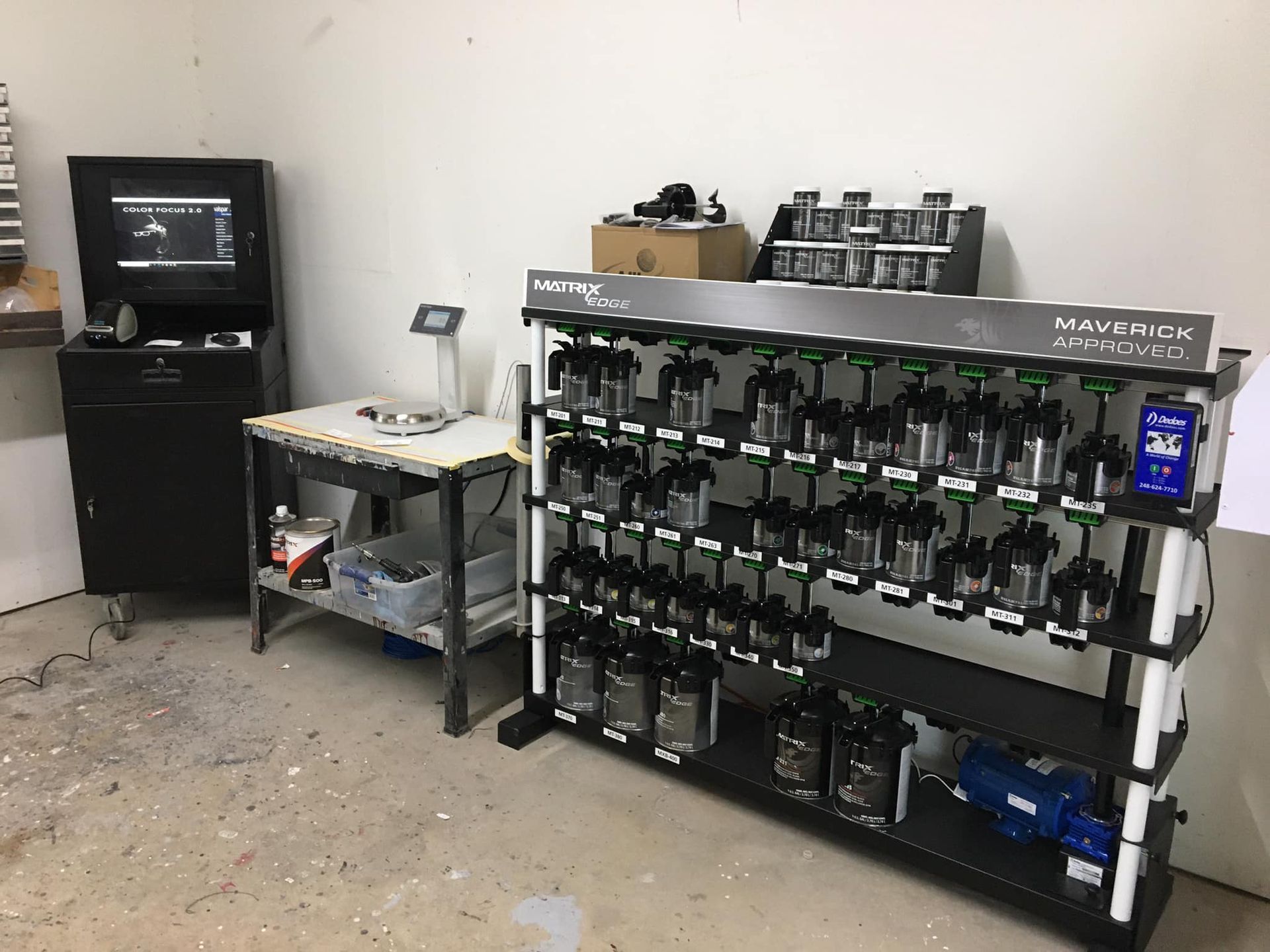 Paint mixing station: multiple paint containers organized on a rack, computer, and a workbench in a workshop.