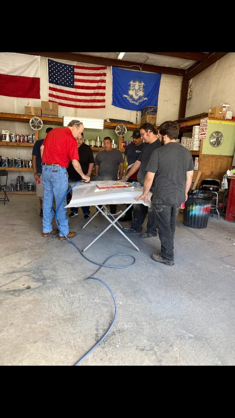 Group of people around a table in a garage. Flags hang in the background. Man in red shirt points at the table.